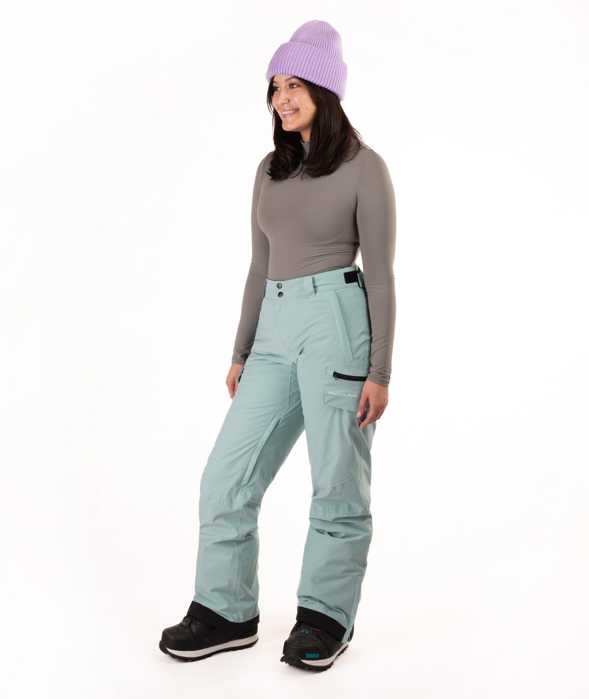 A woman modeling snow pants in a studio setting, wearing a gray long-sleeve top, a light purple knit beanie, and black snow boots against a plain white background.