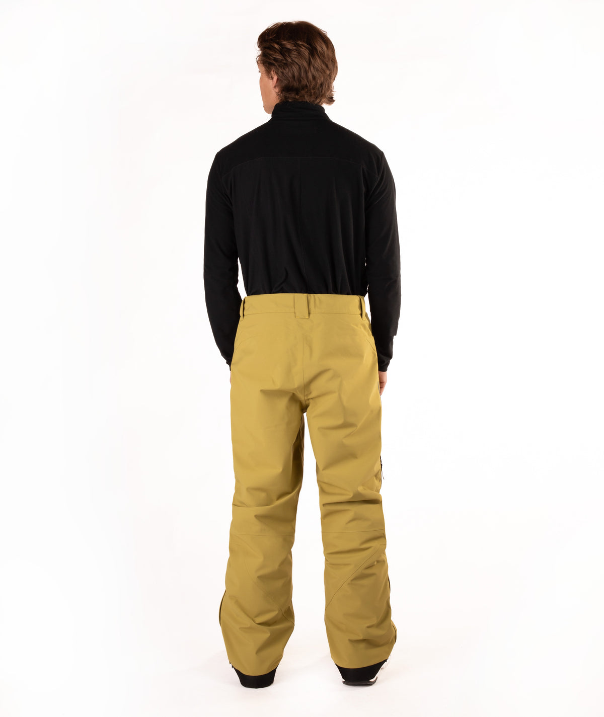 A man viewed from behind wearing a black long-sleeve shirt and mustard yellow snow pants against a white studio background.