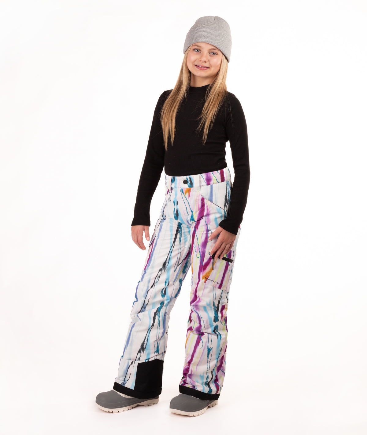 A young girl modeling snow pants with a colorful paint-splatter pattern, paired with a black long-sleeve top, gray beanie, and gray boots, standing against a white studio background.
