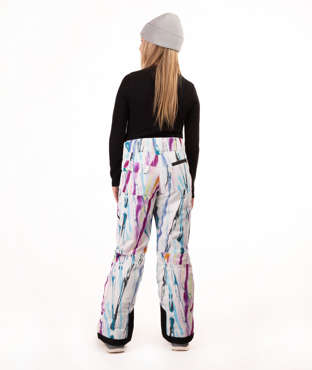 A model shown from the back wearing snow pants with a colorful paint-splatter pattern, a black long-sleeve top, and a gray beanie against a white studio background.