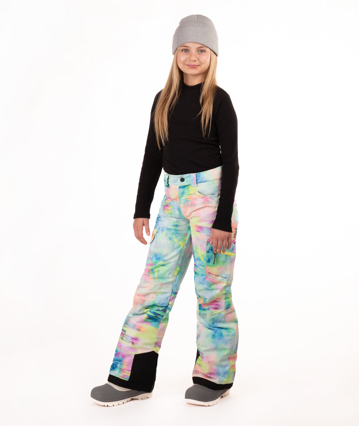 A young girl modeling colorful tie-dye snow pants, a black long-sleeve shirt, and a gray beanie against a white studio background.