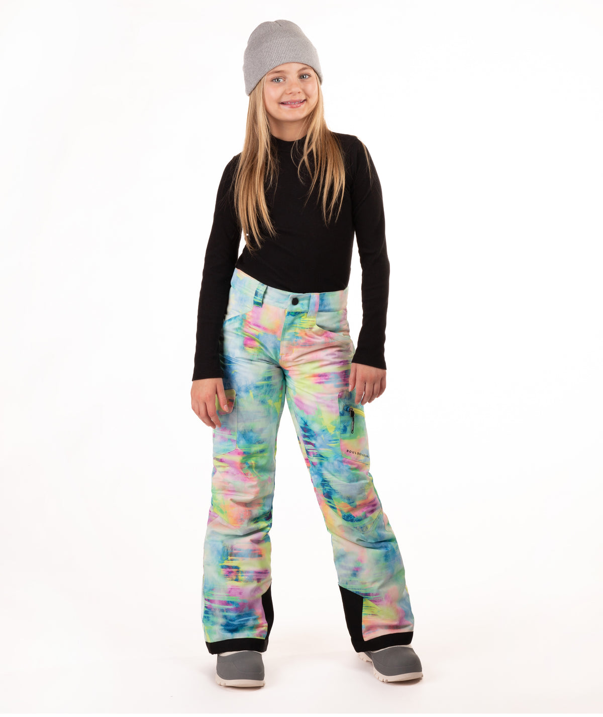 A young girl modeling colorful tie-dye snow pants, a black long-sleeve shirt, and a gray beanie against a white studio background.