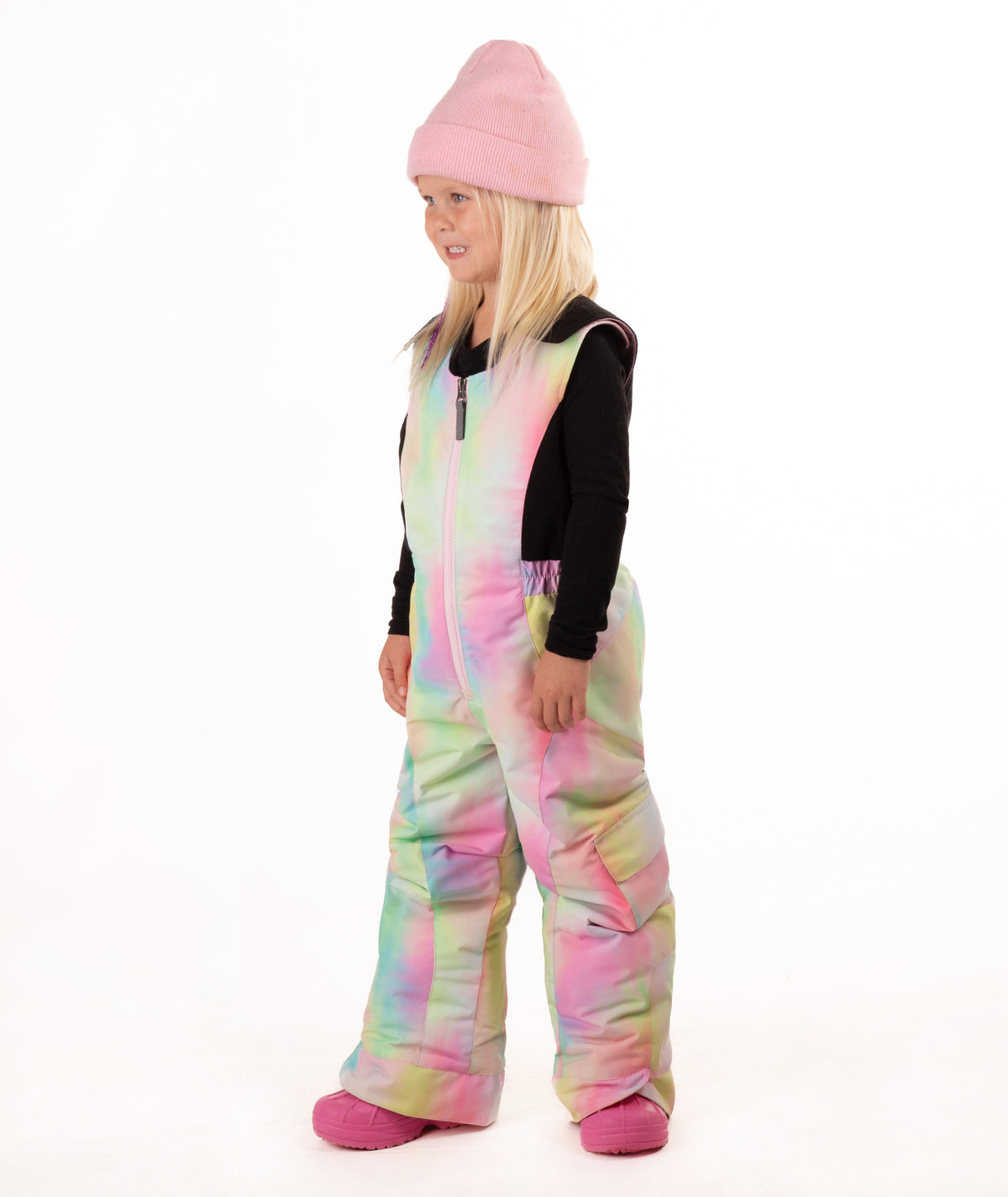 A young girl wearing a pastel rainbow tie-dye snow bib over a black long-sleeve shirt, a pink beanie, and pink boots, standing against a white studio background.