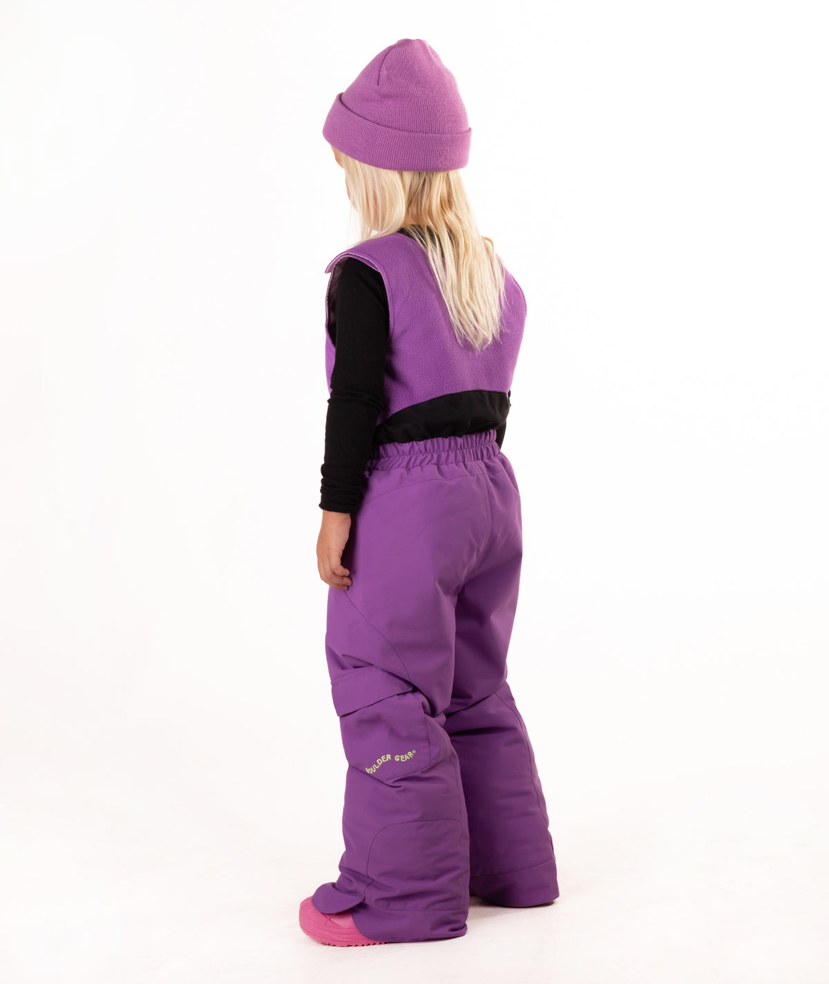 A young child wearing a purple snow bib over a black long-sleeve shirt, a matching purple knit beanie, and pink boots, standing against a plain white studio background.