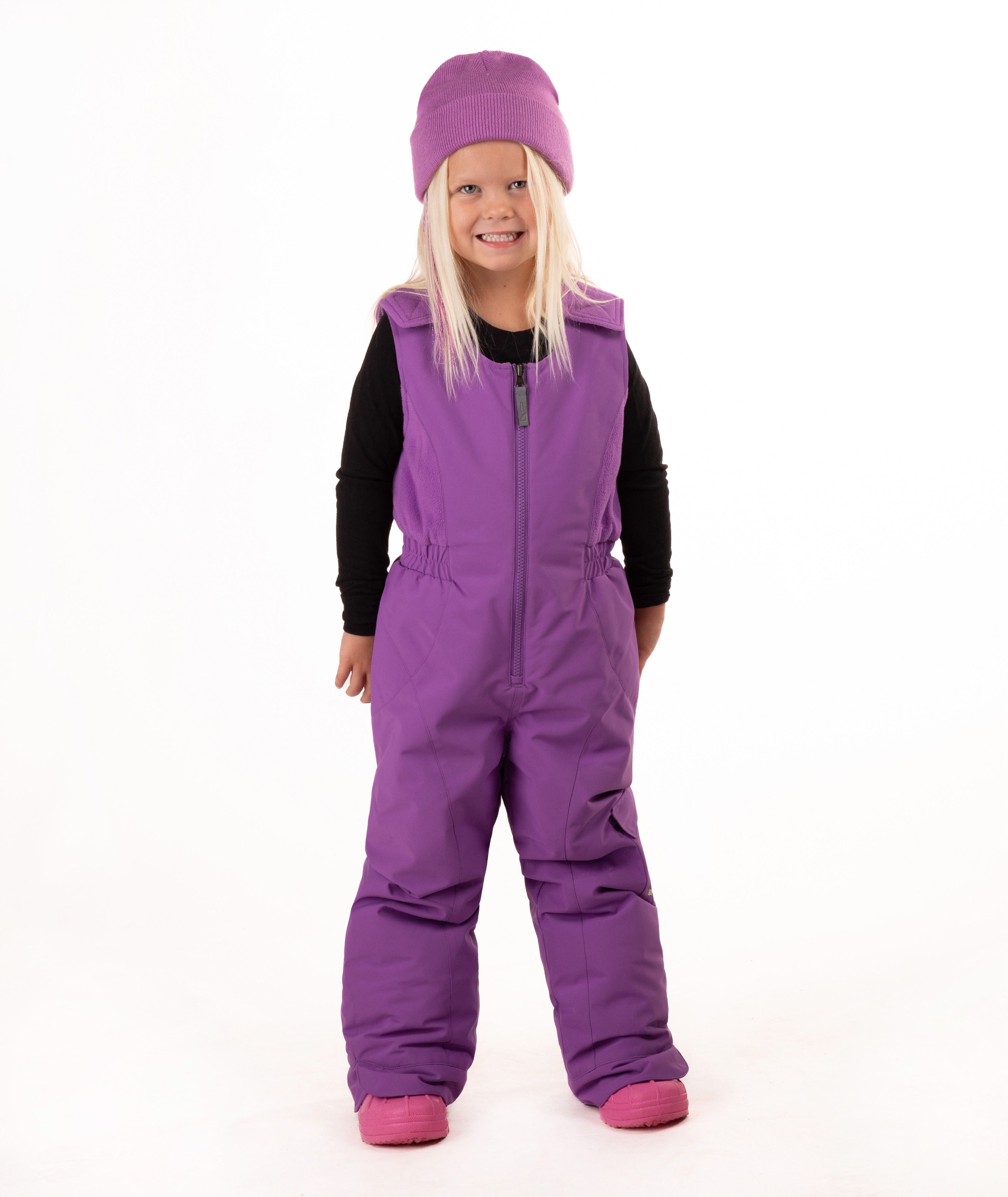 A young child wearing a purple snow bib over a black long-sleeve shirt, a matching purple beanie, and pink boots, standing against a plain white studio background.