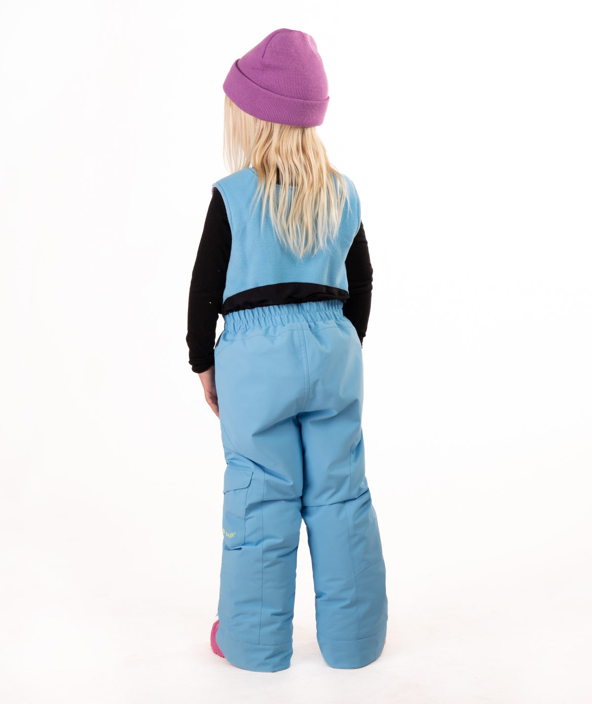 A young child wearing a light blue snow bib over a black long-sleeve shirt, a purple beanie, and pink boots, standing against a plain white studio background.