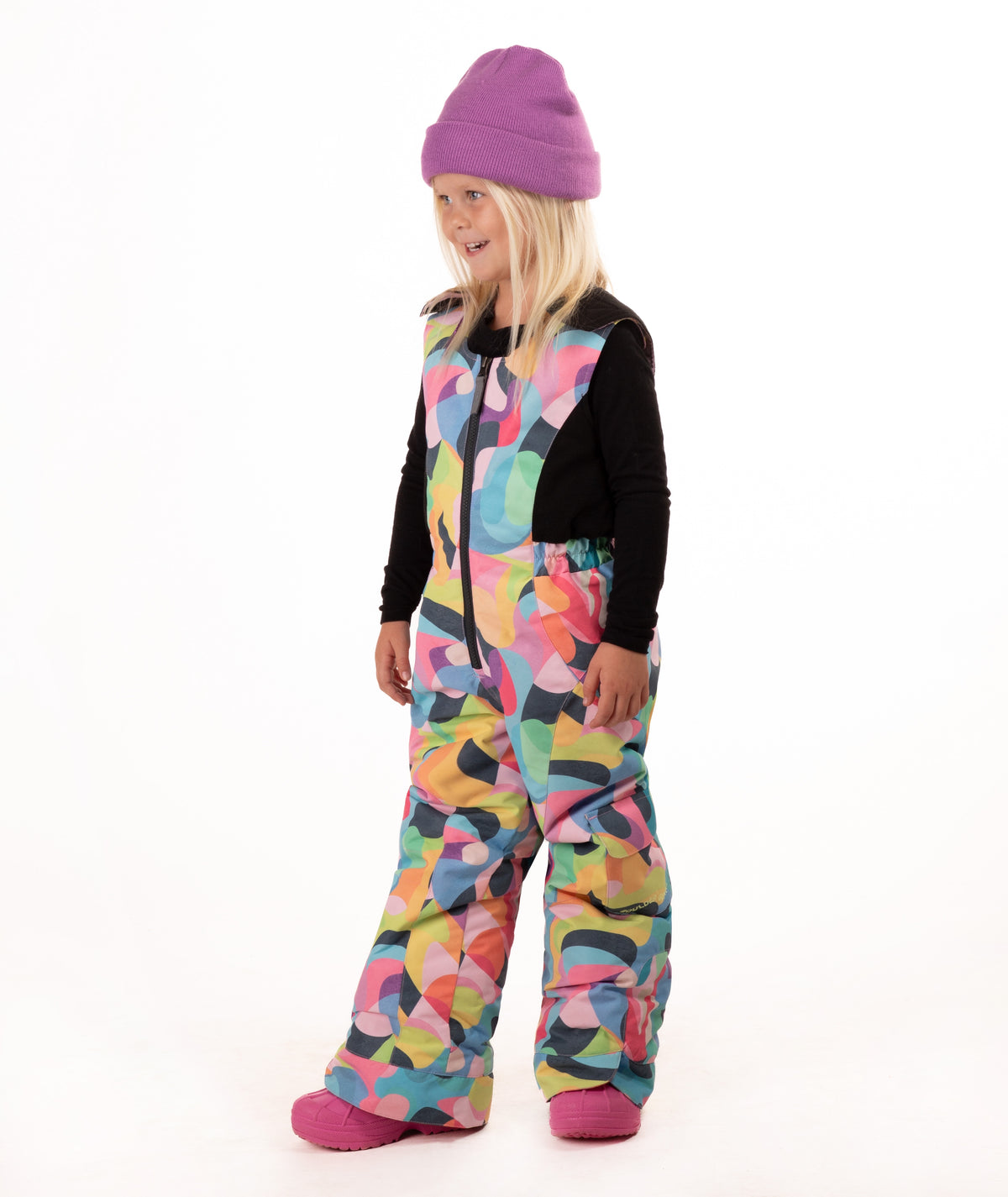 A young child wearing a black long-sleeve top and a colorful abstract print snowsuit with a purple beanie and pink boots, standing against a white studio background.
