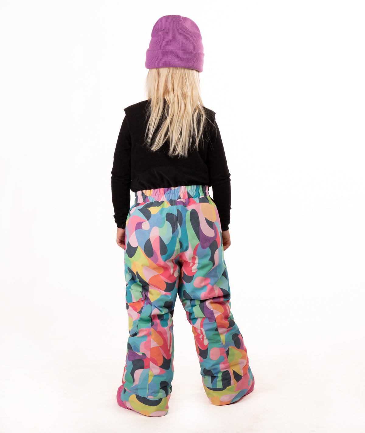 A young child wearing a black long-sleeve top and a colorful abstract print snowsuit with a purple beanie and pink boots, standing against a white studio background.