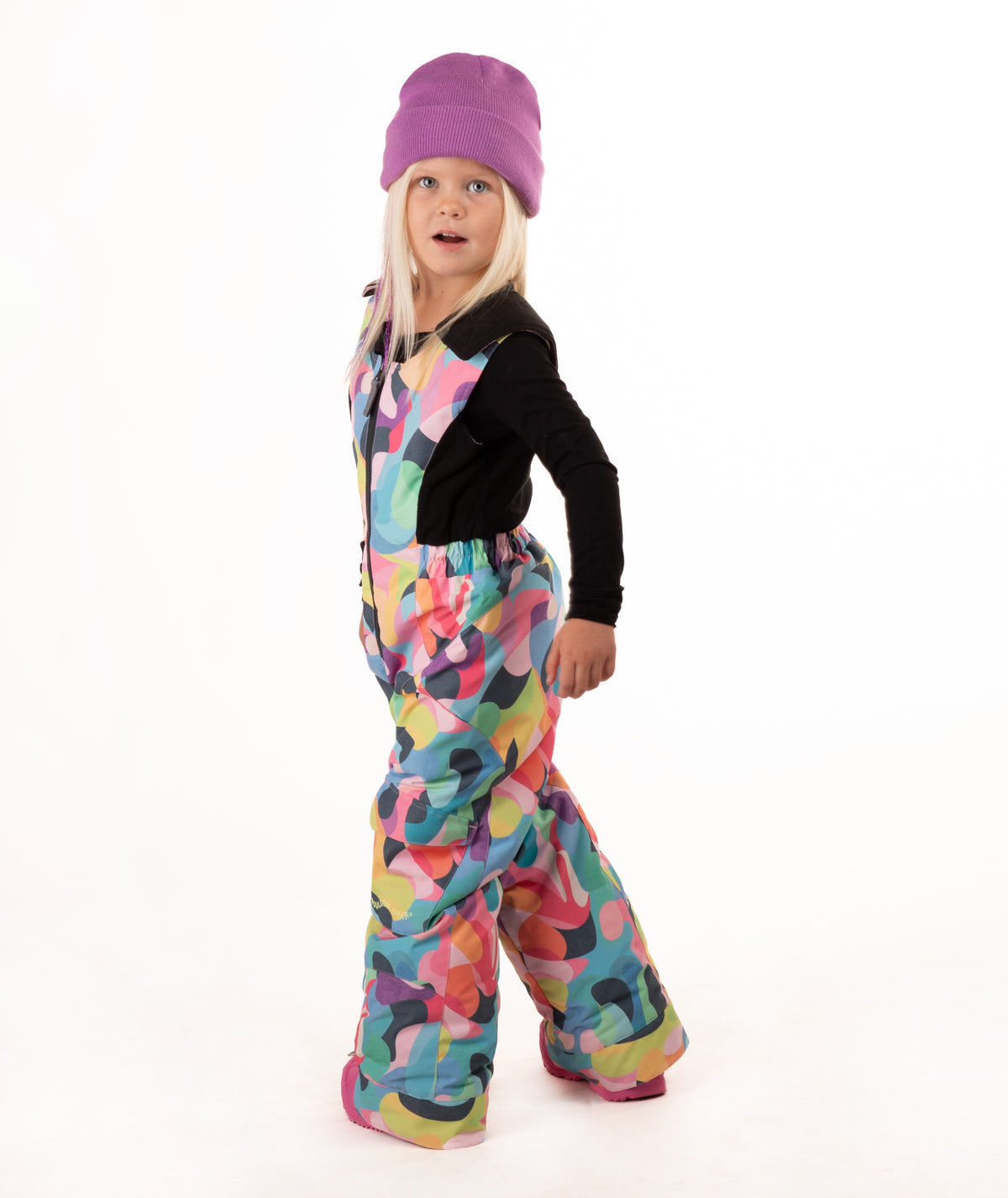 A young child wearing a black long-sleeve top and a colorful abstract print snowsuit with a purple beanie and pink boots, standing against a white studio background.