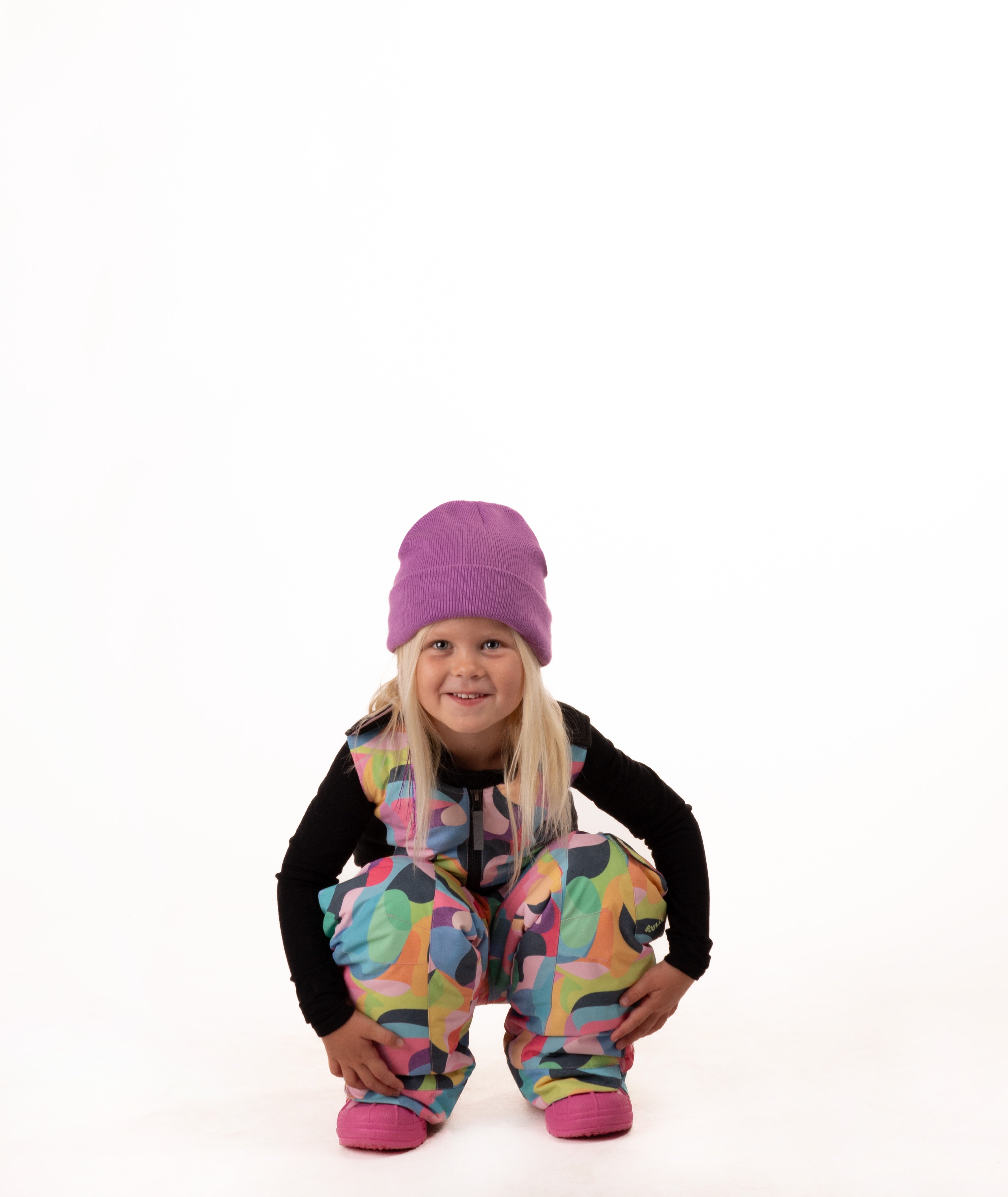 A young girl crouching down in colorful snow pants and a purple beanie, wearing a black long-sleeve shirt, against a solid white background.