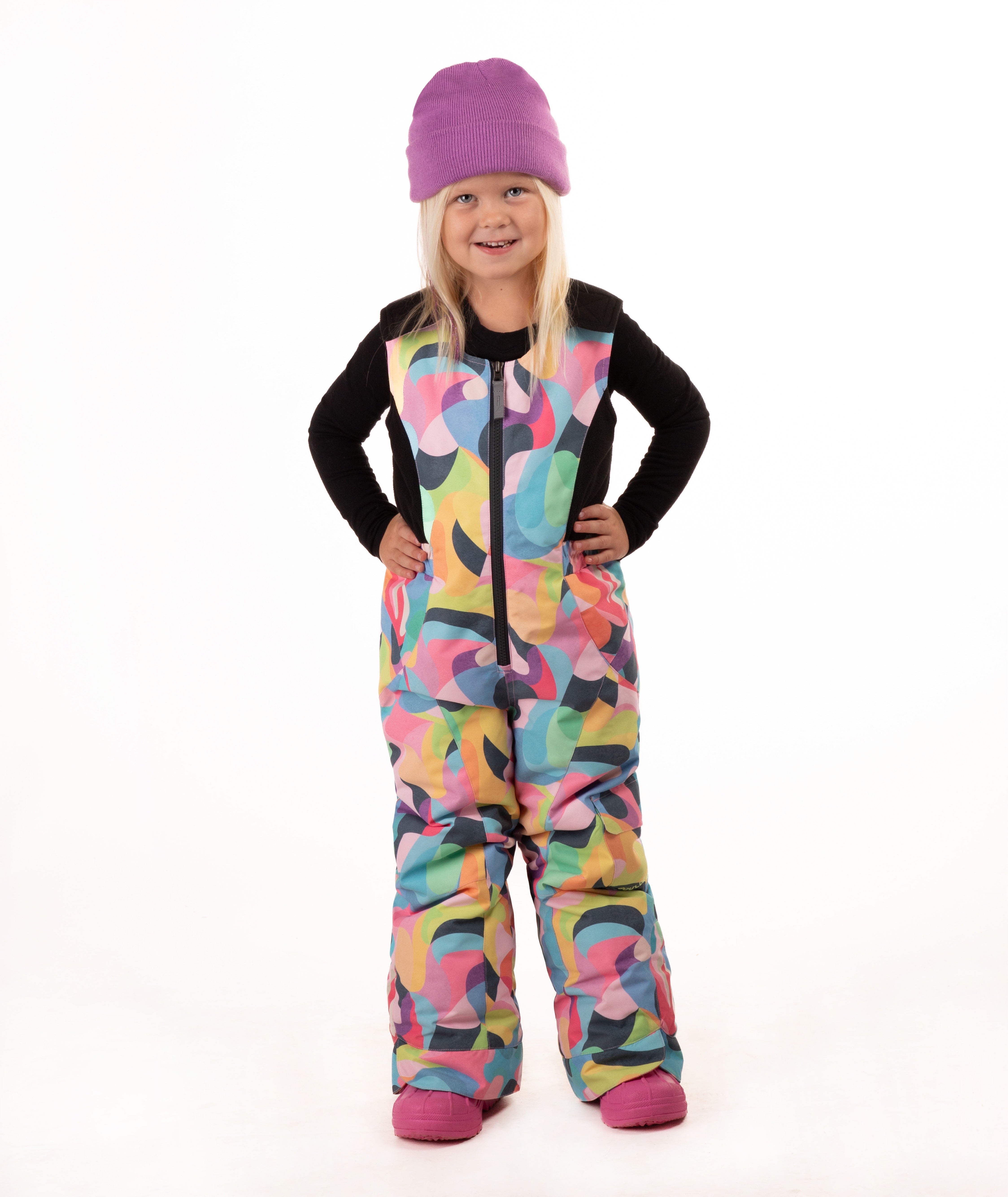 A young child wearing a black long-sleeve top and a colorful abstract print snowsuit with a purple beanie and pink boots, standing against a white studio background.
