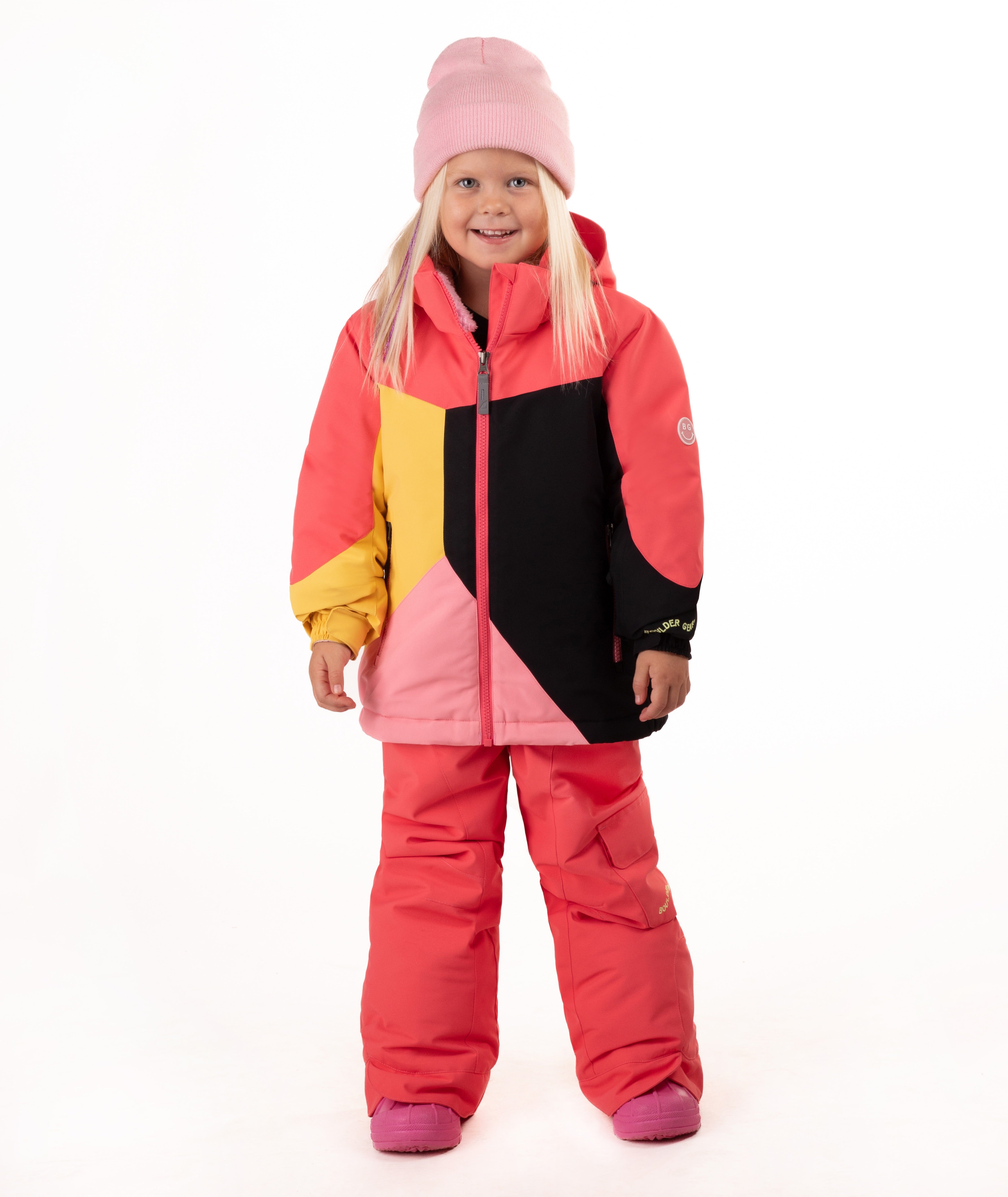 A young girl wearing a color-blocked ski jacket in pink, coral, yellow, and black, matching snow pants, a pink beanie, standing against a white studio background.