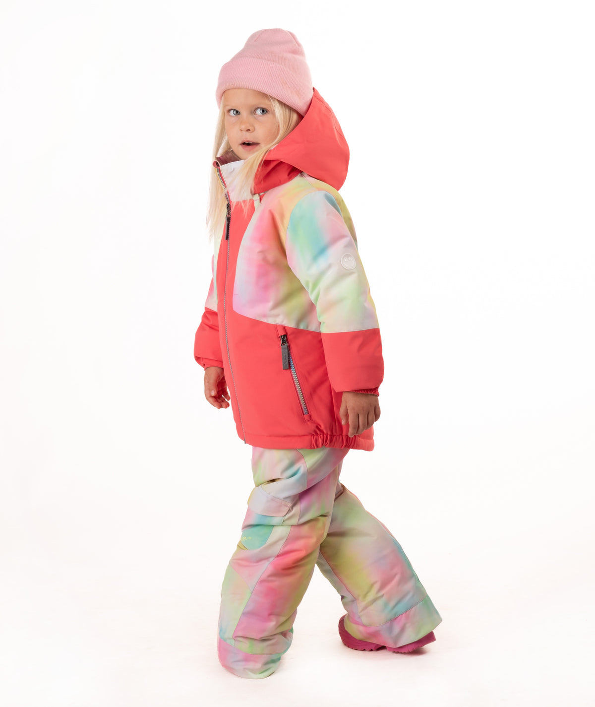 A young girl wearing a pink tie-dye jacket, consisting of a bright pink jacket and matching pants, along with a pink beanie and pink boots, standing against a white studio background.