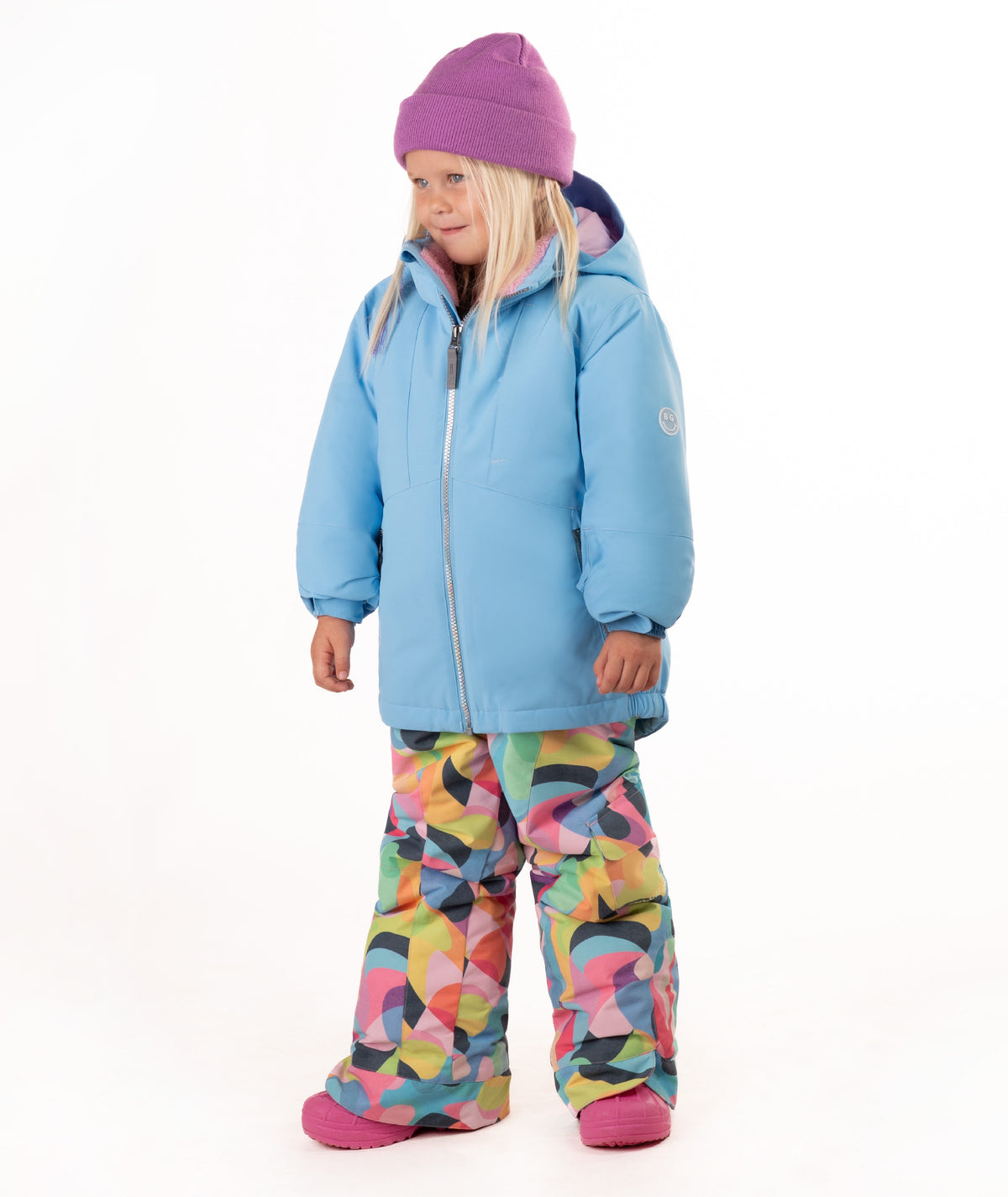 A young child wearing a light blue winter jacket, a purple beanie, and colorful geometric print snow pants, standing against a plain white background.