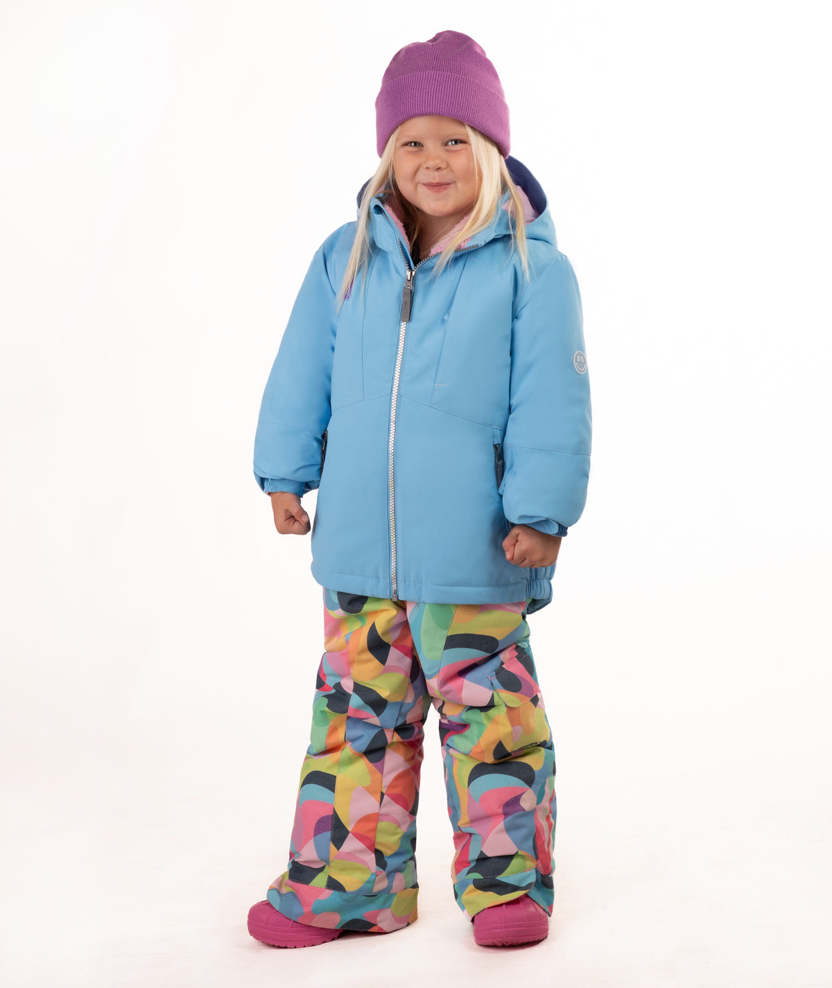 A young child wearing a light blue winter jacket, a purple beanie, and colorful geometric print snow pants, standing against a plain white background.