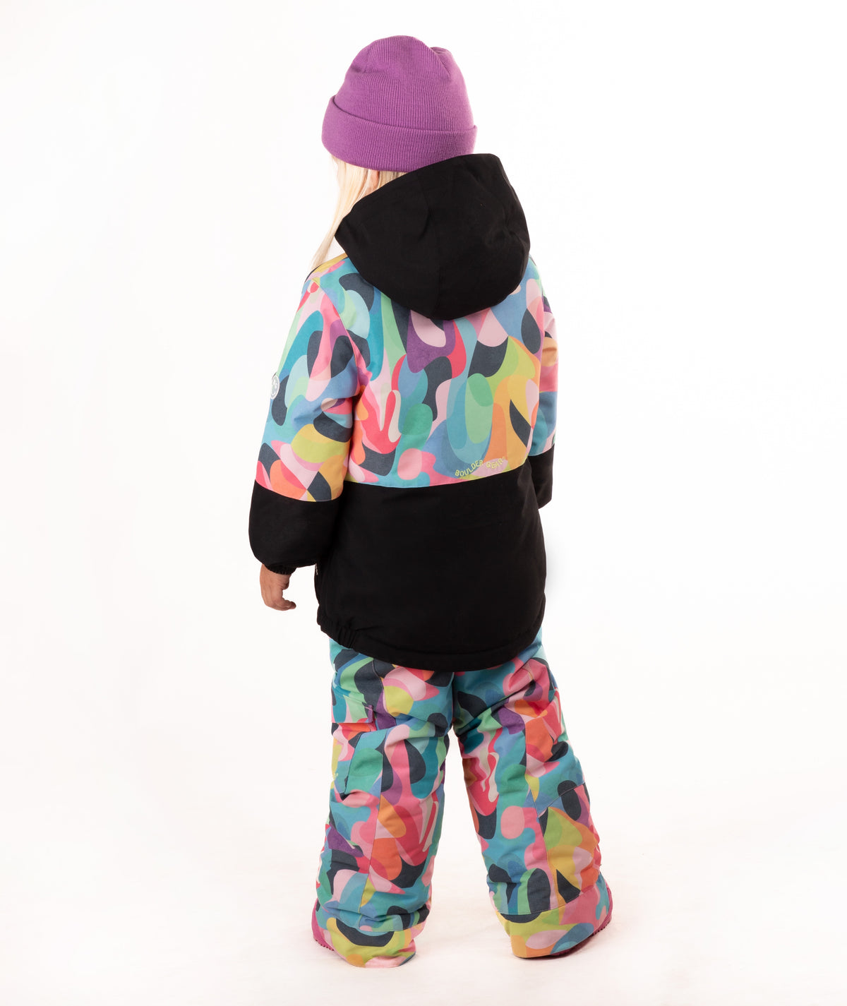 A young girl wearing a colorful abstract print ski jacket and matching pants, a purple beanie, and pink boots, standing against a white studio background.