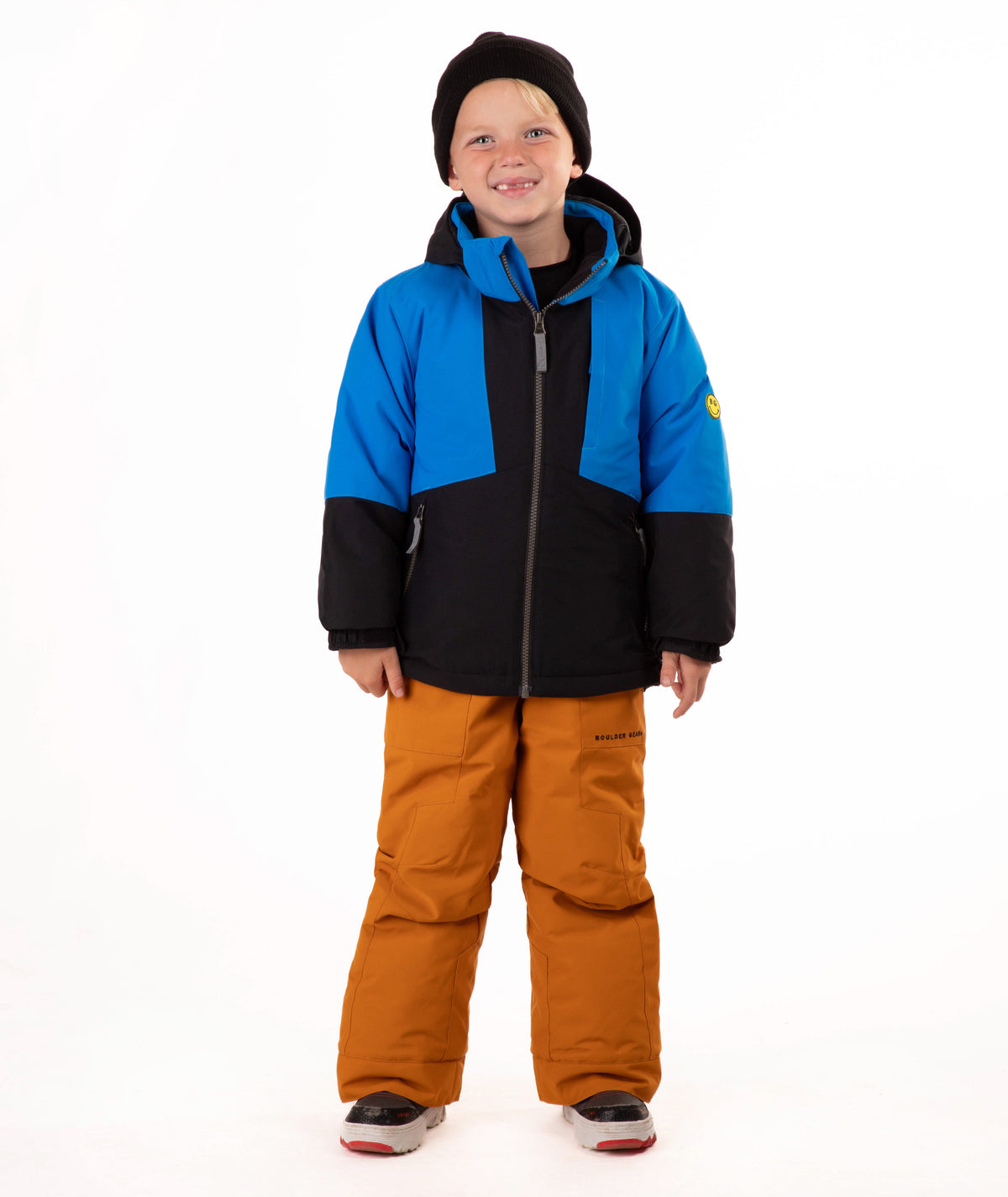 A child standing against a white studio background wearing a color-blocked ski jacket in blue and black, brown snow pants, and a black beanie