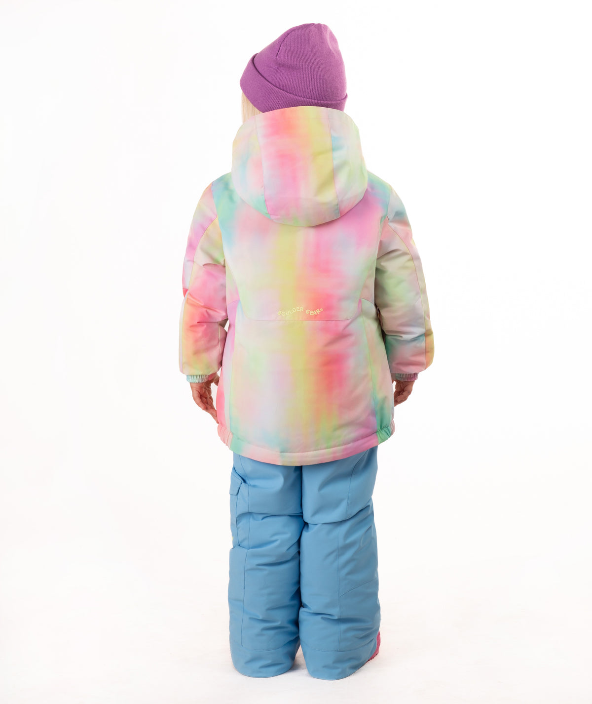 A young girl stands against a white studio background wearing a pastel tie-dye hooded snow jacket, light blue snow pants, and a purple beanie.
