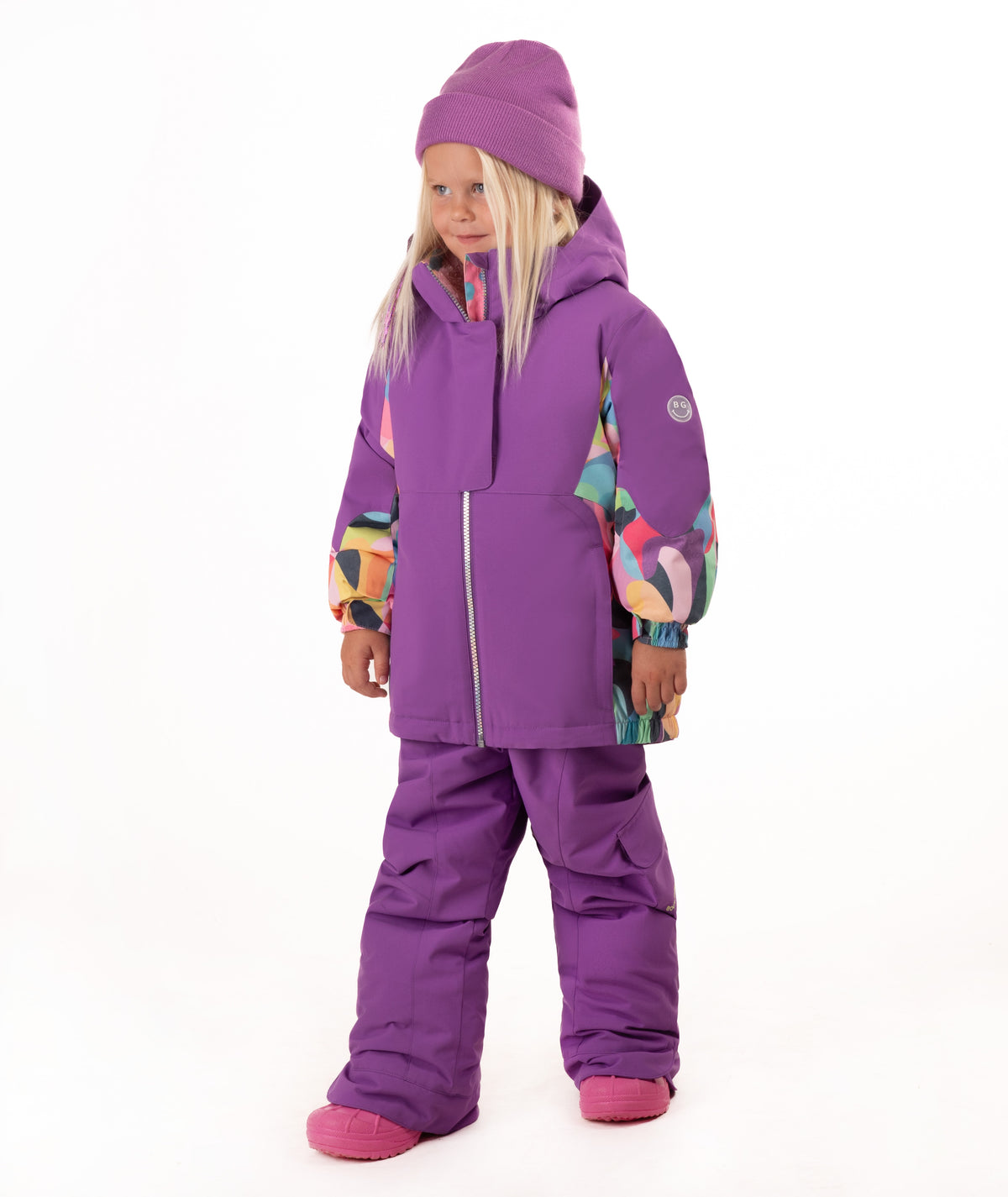 A young girl wearing a purple ski outfit with colorful geometric patterned accents on the sleeves, a matching purple beanie, and purple snow pants, standing against a white background.