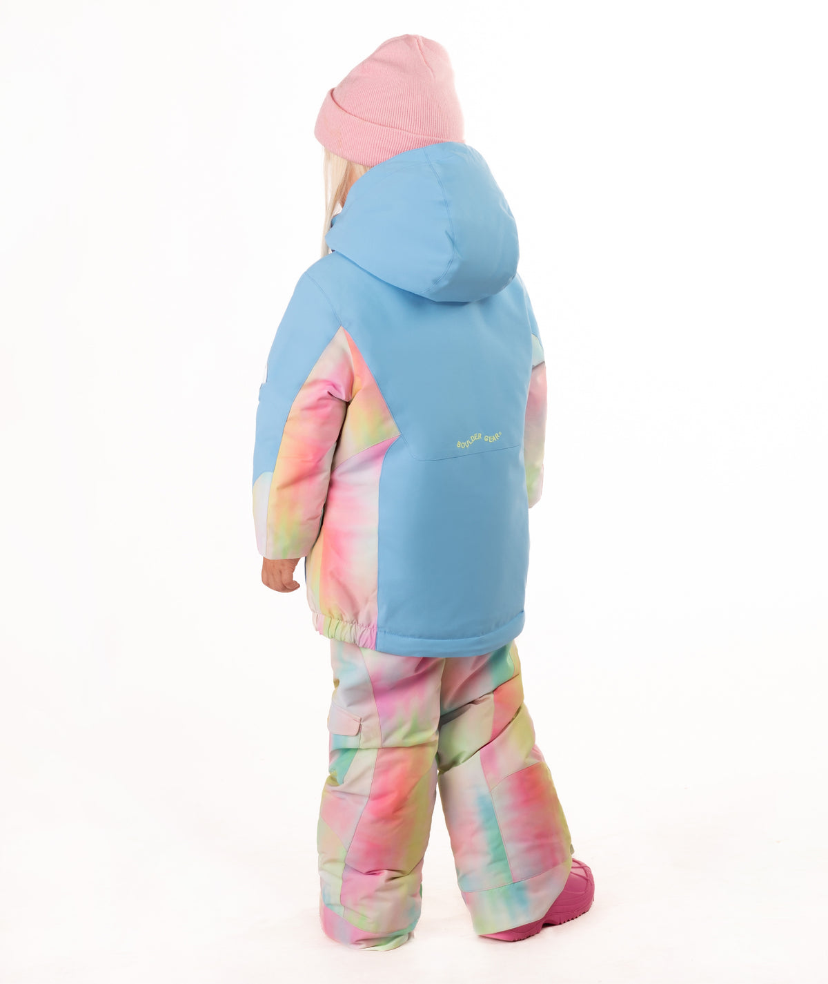 A young child wearing a light blue ski jacket with pastel tie-dye panels, matching tie-dye snow pants, a pink beanie, and pink boots, standing against a plain white background.
