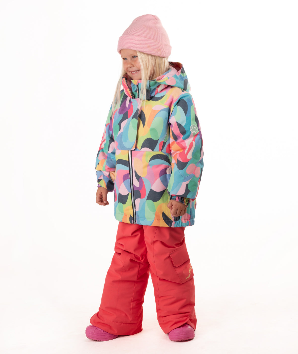 A young child wearing a colorful abstract-patterned ski jacket, bright pink snow pants, and a pink beanie, standing against a plain white studio background.