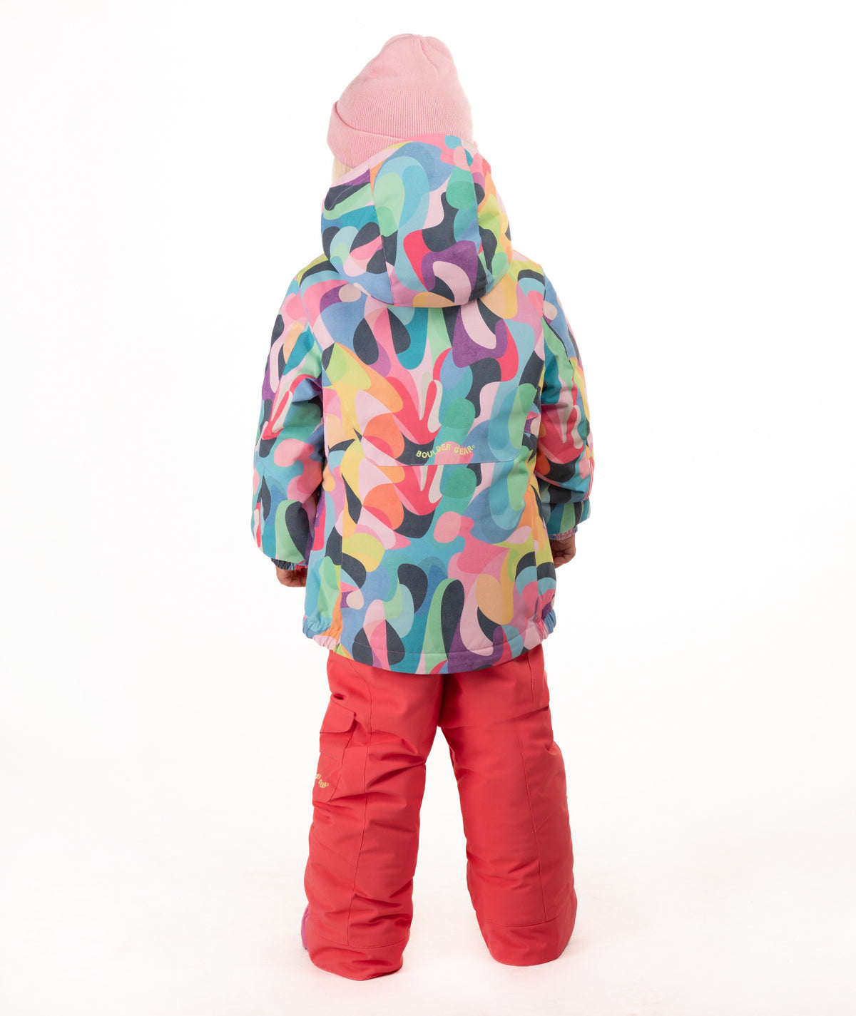 A young child wearing a colorful abstract-patterned ski jacket, bright pink snow pants, and a pink beanie, standing against a plain white studio background.