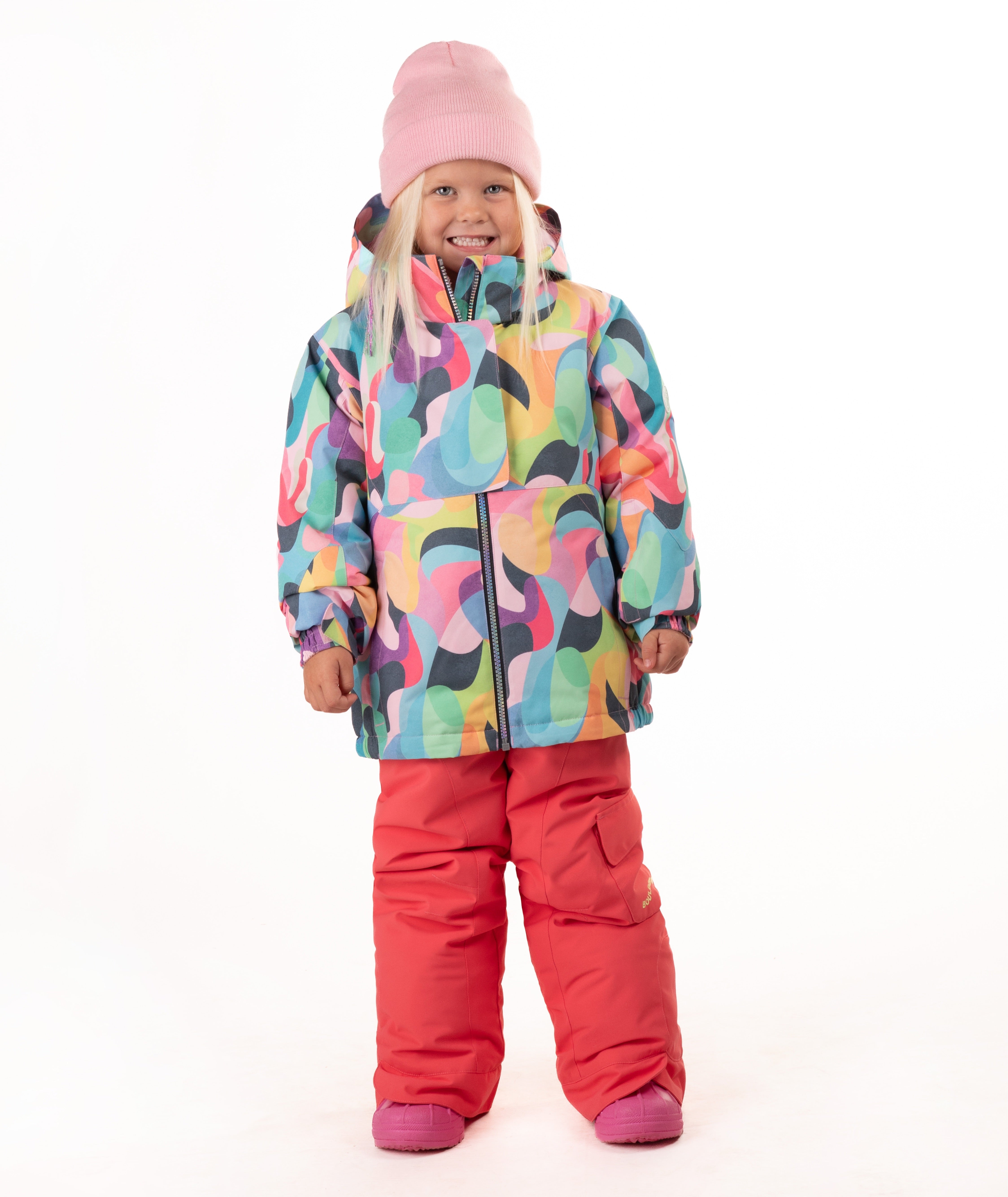 A young child wearing a colorful abstract-patterned ski jacket, bright pink snow pants, and a pink beanie, standing against a plain white studio background.