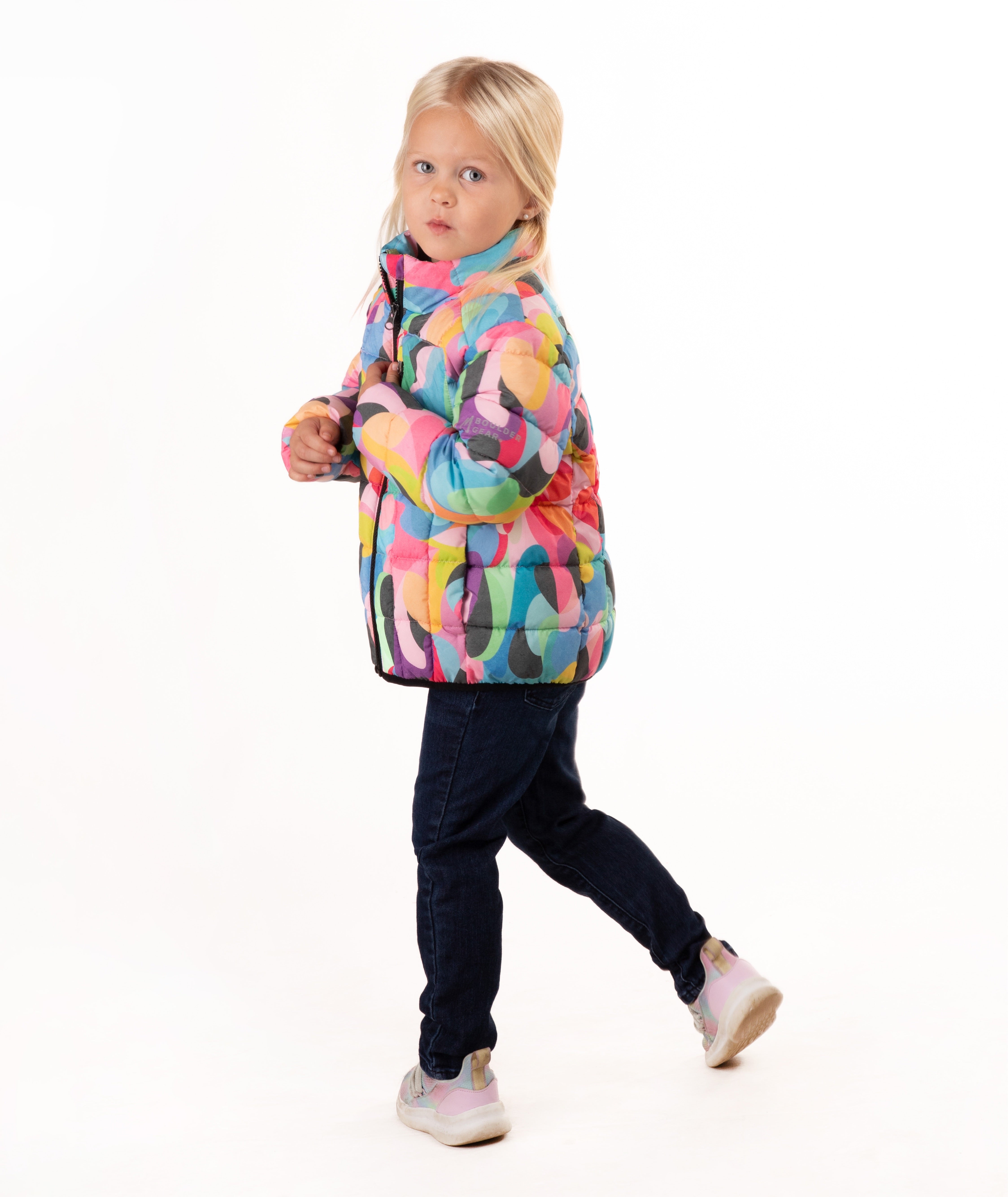 A young girl with blonde hair stands against a white studio background wearing a colorful abstract print puffer jacket, dark blue jeans, and light gray sneakers.