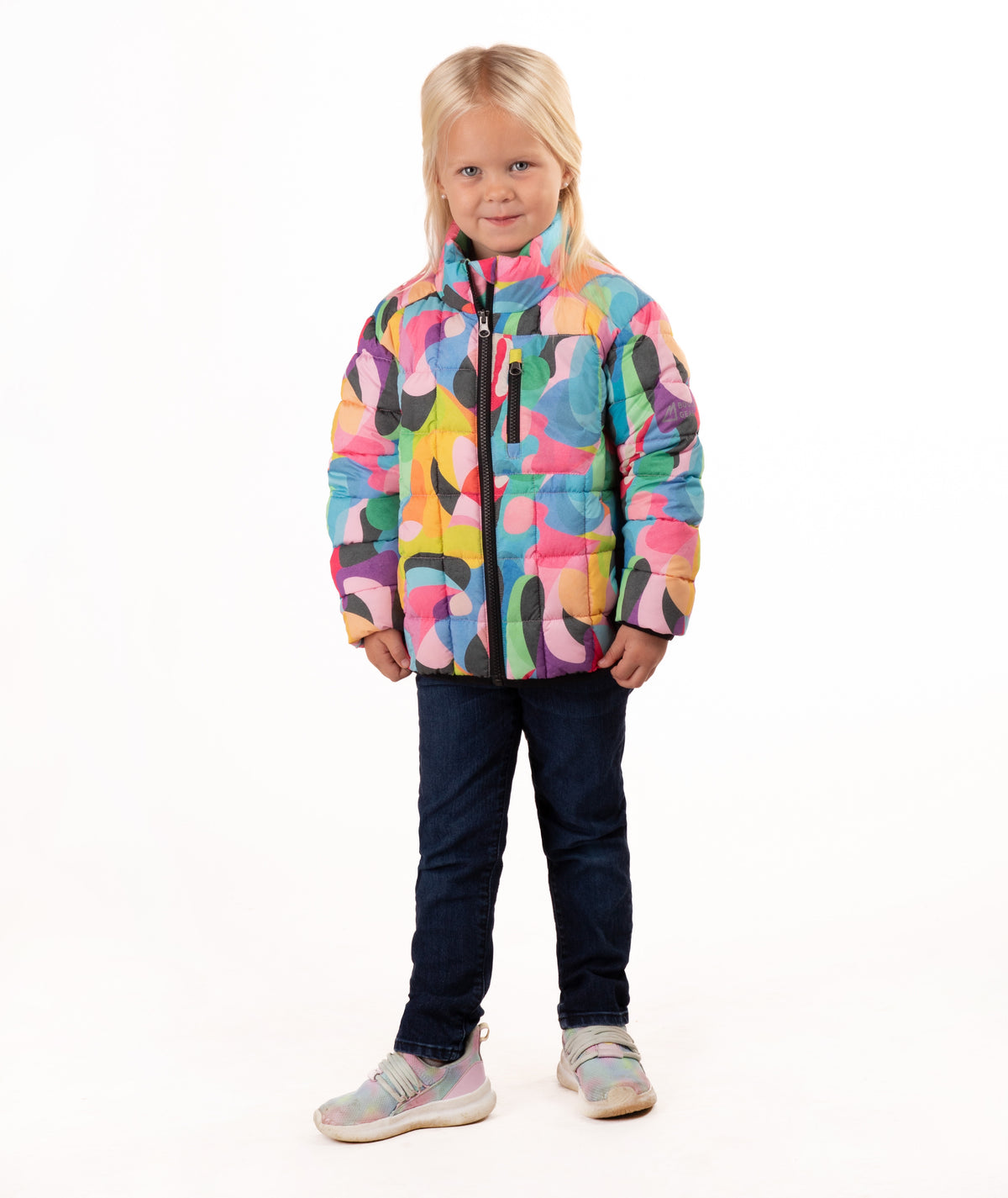 A young girl with blonde hair stands against a white studio background wearing a colorful abstract print puffer jacket, dark blue jeans, and light gray sneakers.