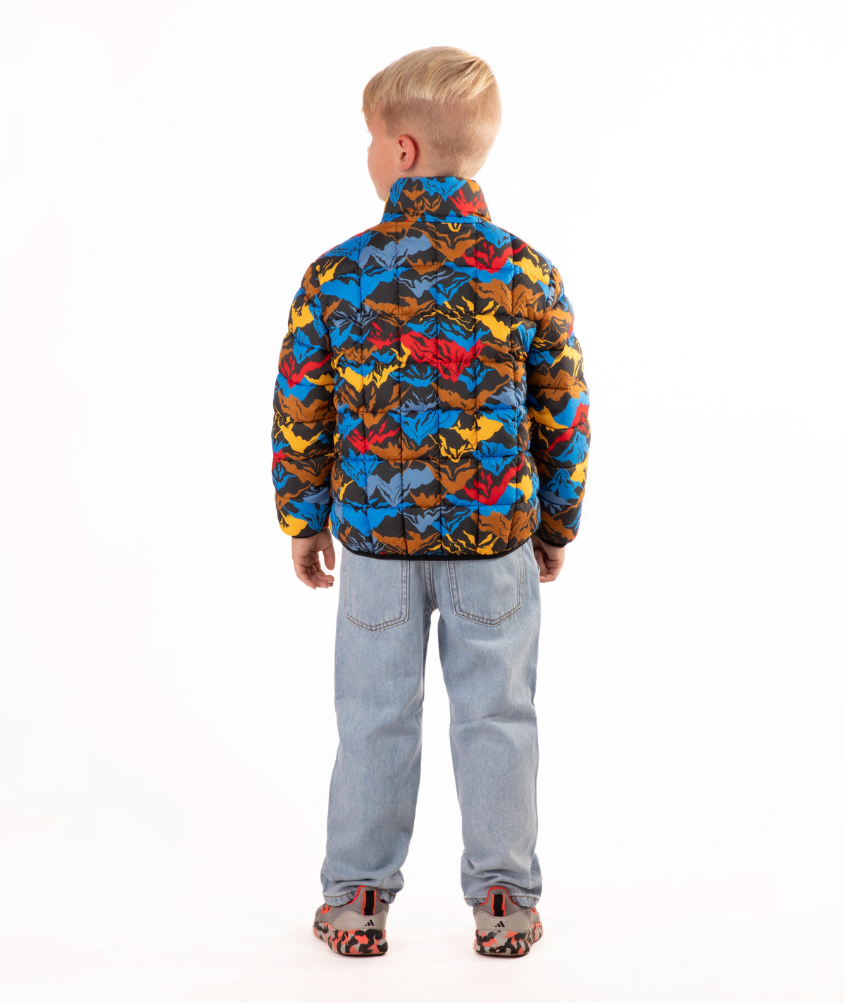 A young boy stands against a white studio background wearing a colorful puffer jacket with a multi color mountain print, blue jeans, and brown shoes.