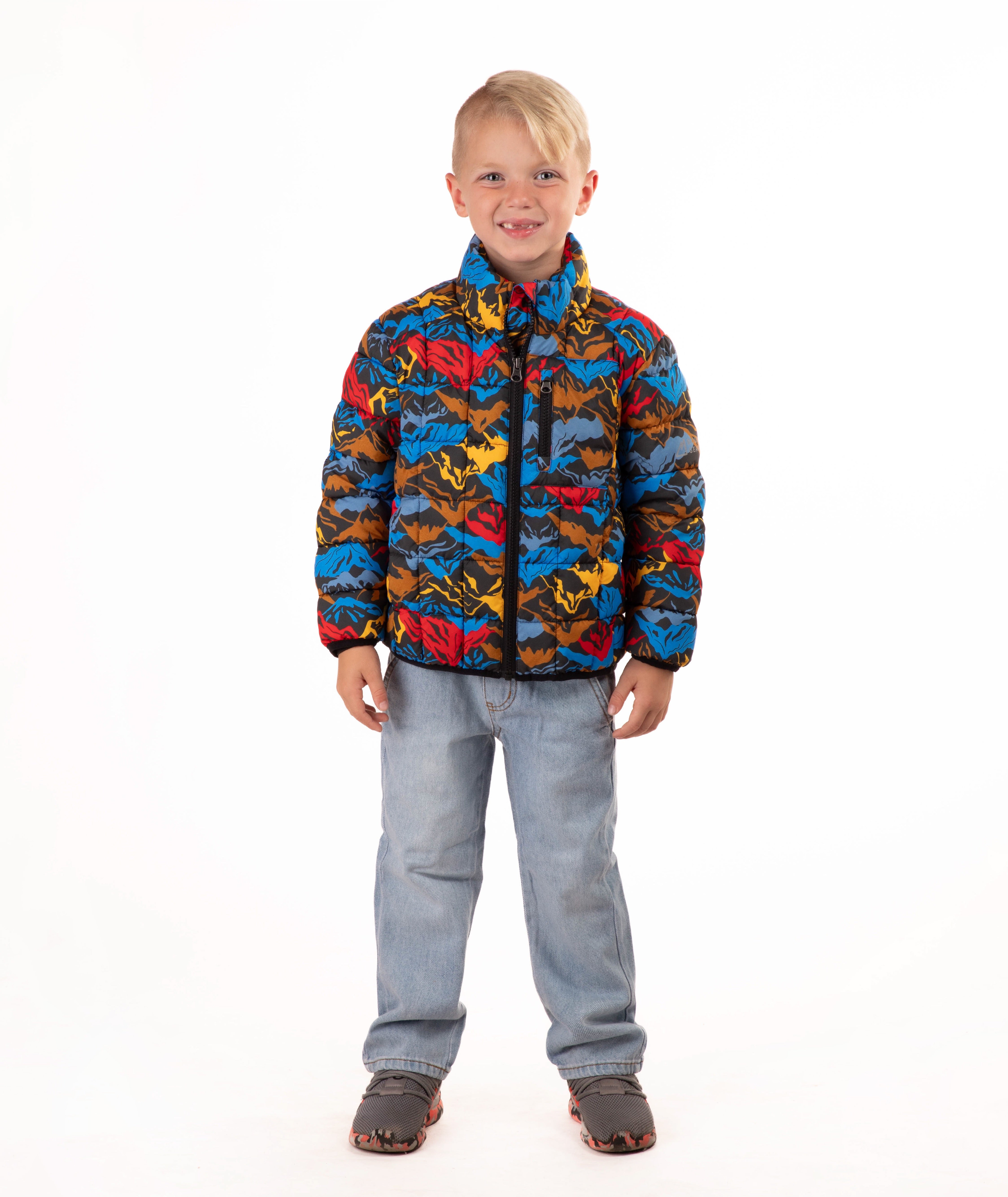 A young boy stands against a white studio background wearing a colorful puffer jacket with a multi color mountain print, blue jeans, and brown shoes.