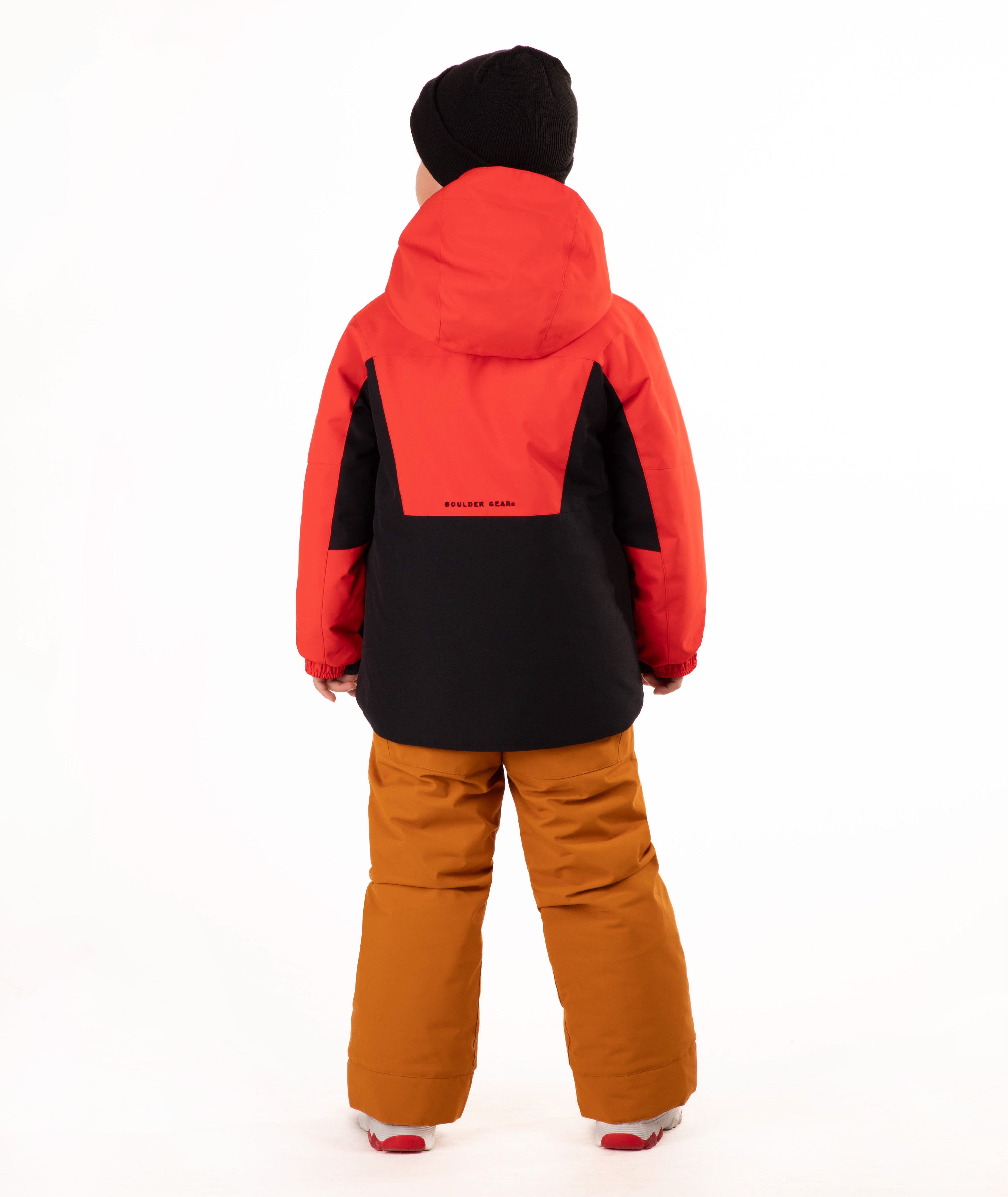 A young child wearing a red and black hooded snow jacket, rust-orange snow pants, a black beanie, standing against a plain white studio background.