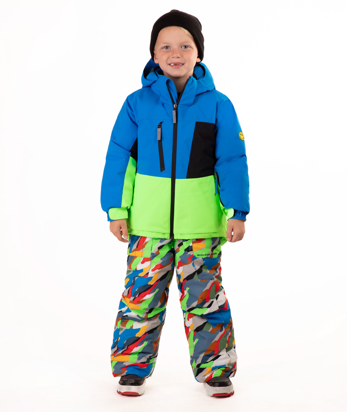 A child wearing a bright blue and green ski jacket with a black chest panel, colorful camouflage-patterned snow pants, and a black beanie, standing against a plain white background.