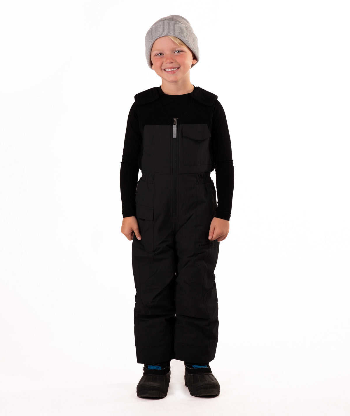A child wearing black snow bib overalls over a black long-sleeve shirt and black beanie, standing against a plain white studio background.