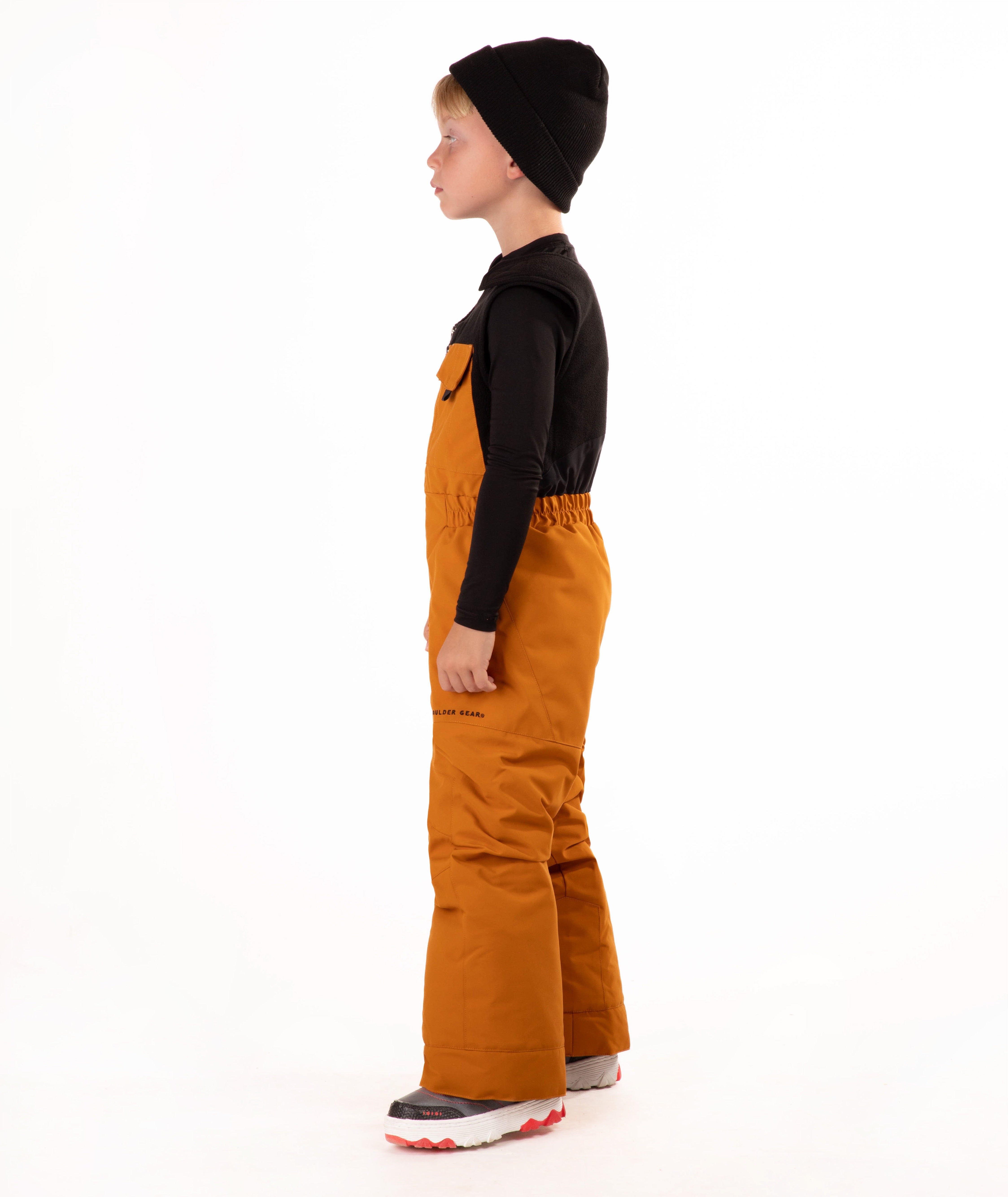 A child wearing a black knit beanie, a black long-sleeve top, and rust colored snow bib overalls, standing against a plain white background.