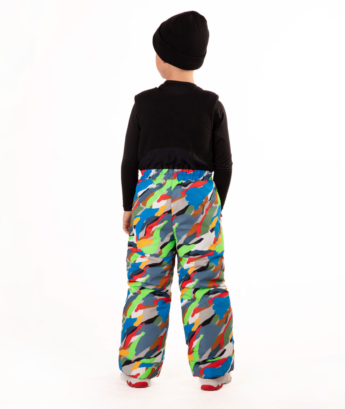 A child wearing a black knit beanie, a black long-sleeve top, and colorful camo print snow bib overalls, standing against a plain white background.