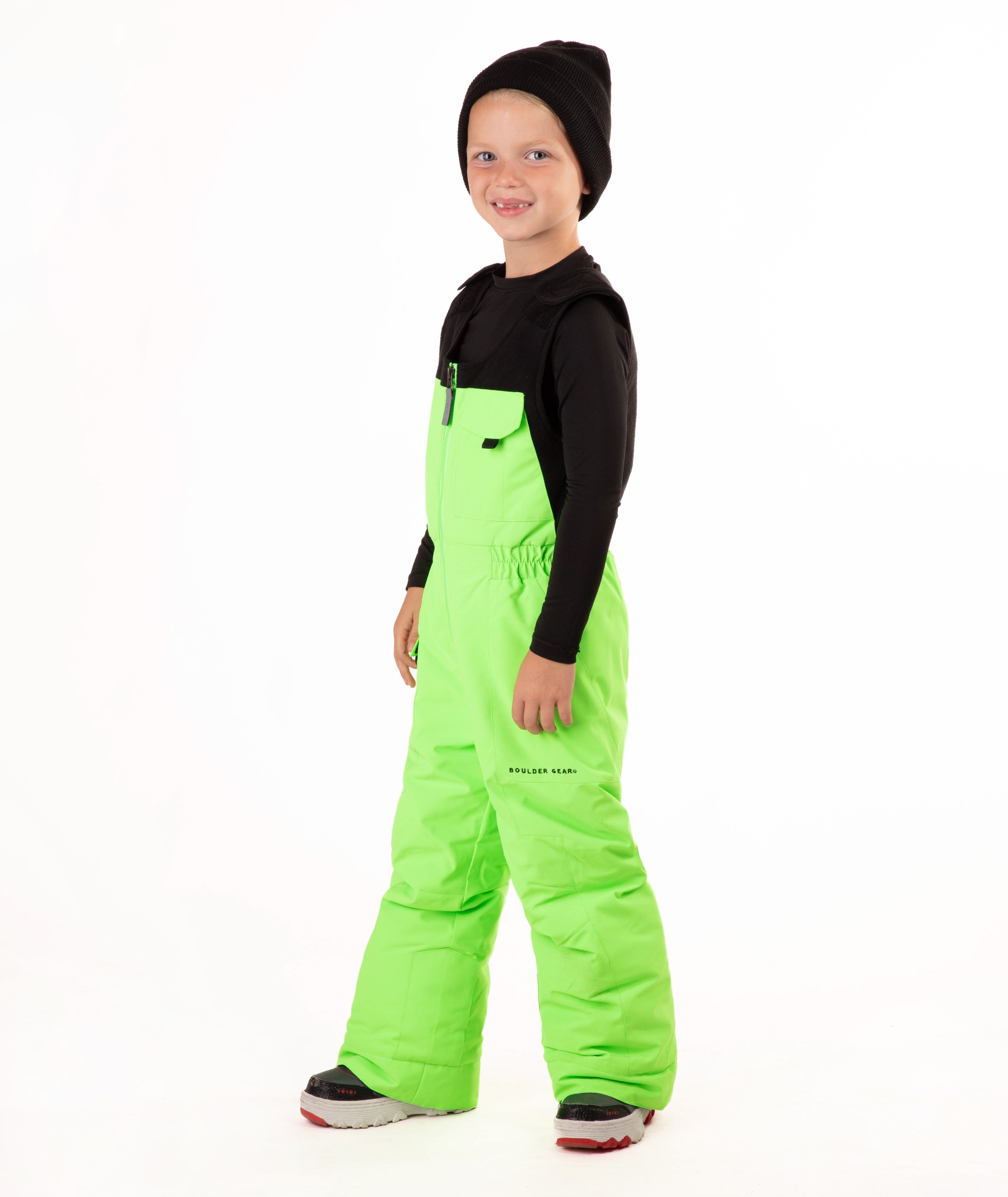 A child wearing bright neon green snow bib overalls over a black long-sleeve shirt and black beanie, standing against a plain white studio background.