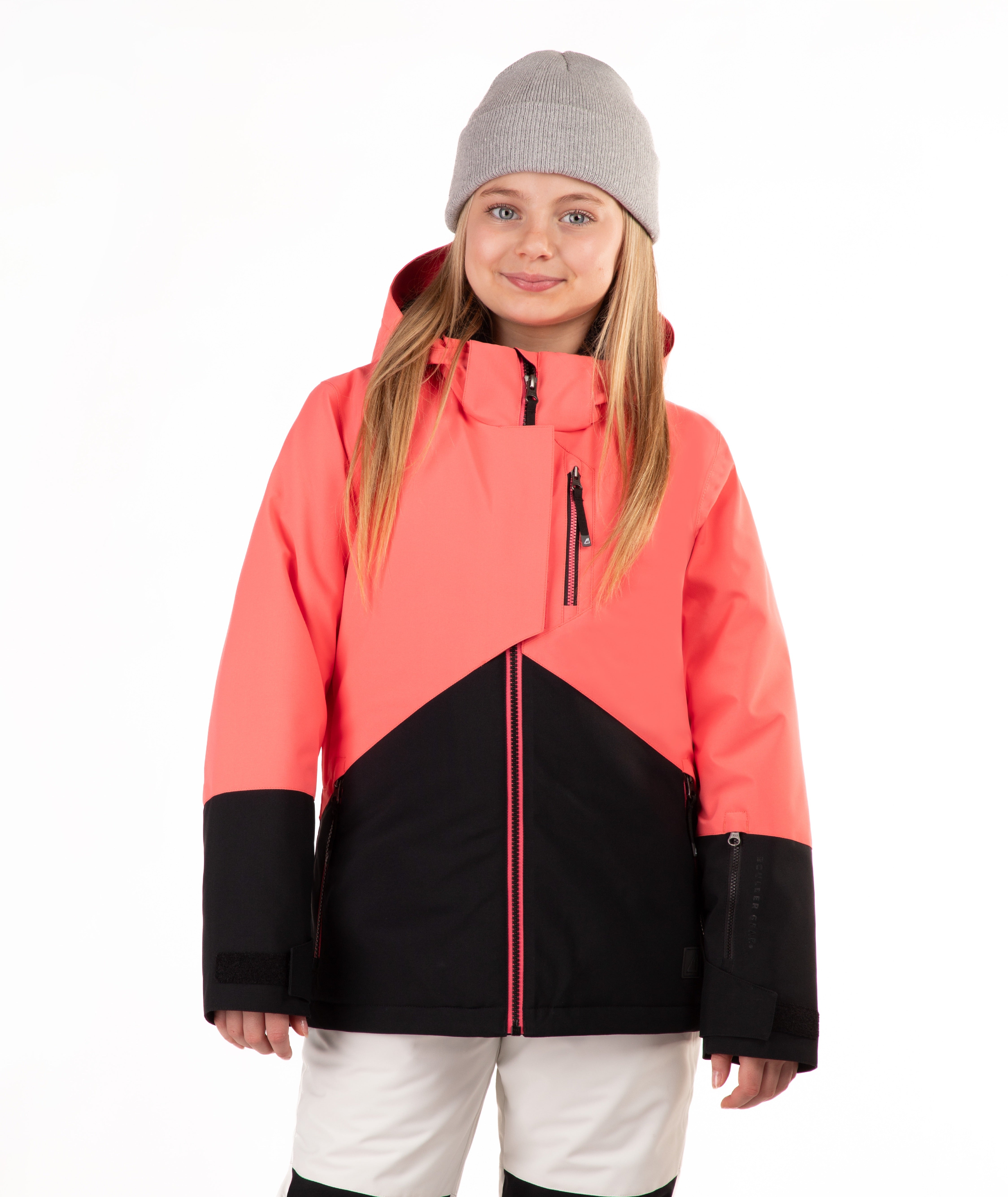 A young girl wearing a coral pink and black color-blocked ski jacket, a gray beanie, and off white ski pants, standing against a solid white background.