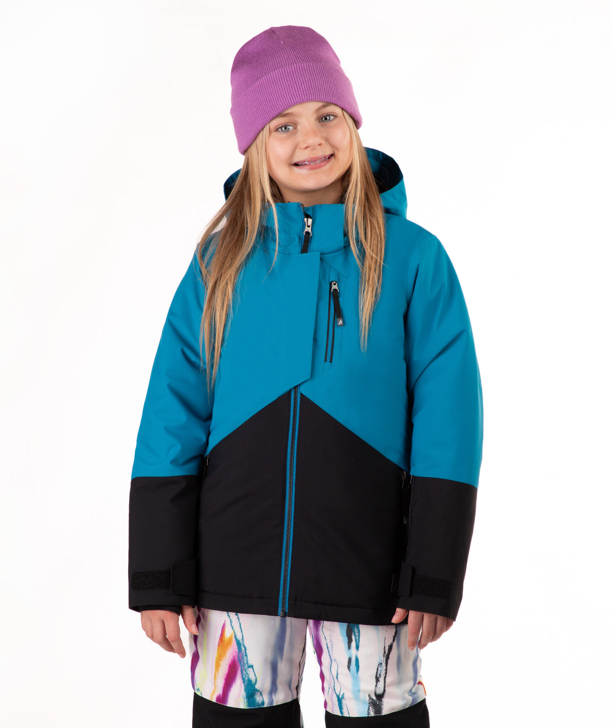 A young girl wearing a colorblock ski jacket in teal and black, a purple beanie, and colorful patterned ski pants, standing against a plain white background.