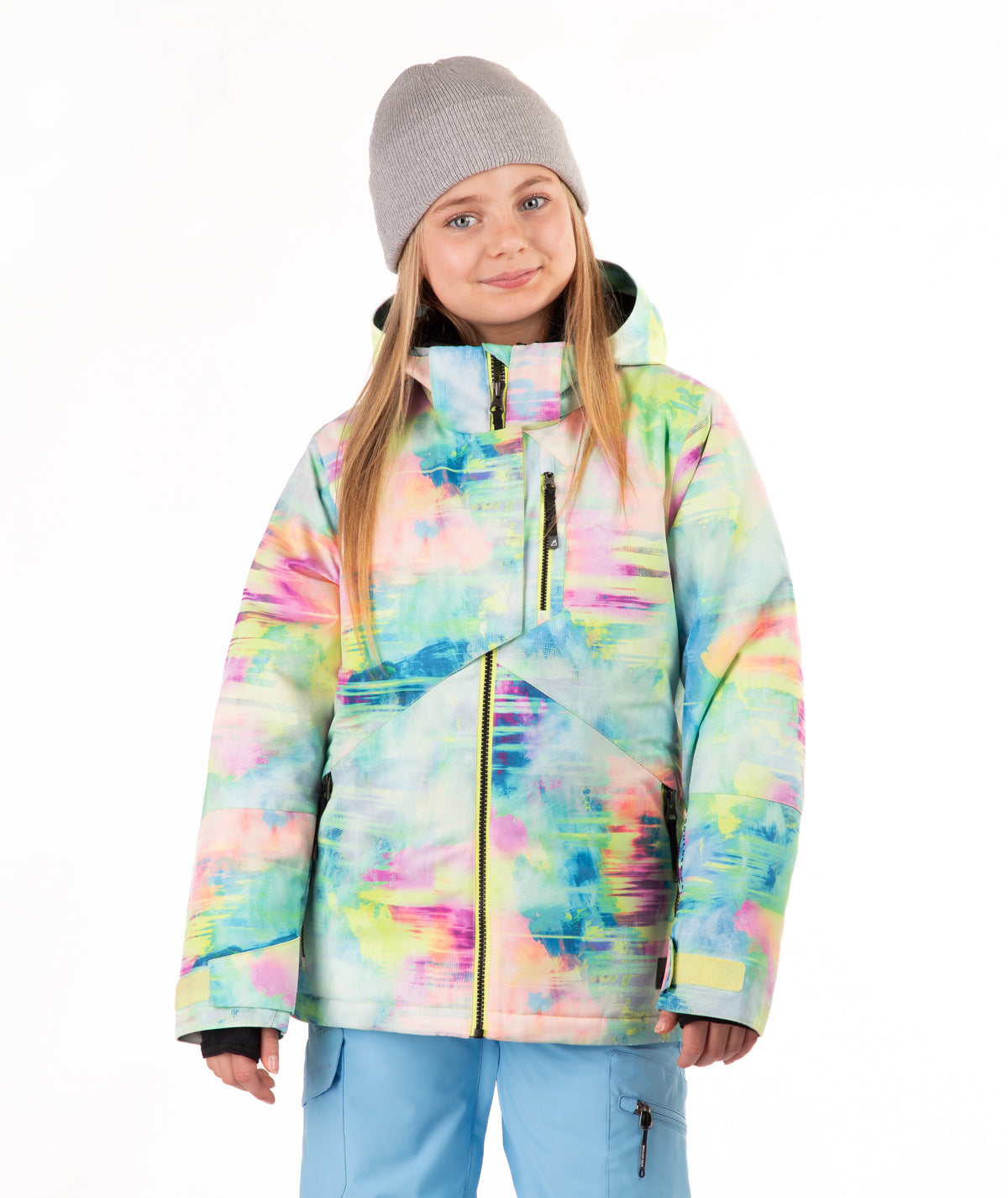 A young girl wearing a colorful tie-dye snowboard jacket, a gray beanie, and light blue snow pants, standing against a plain white background.