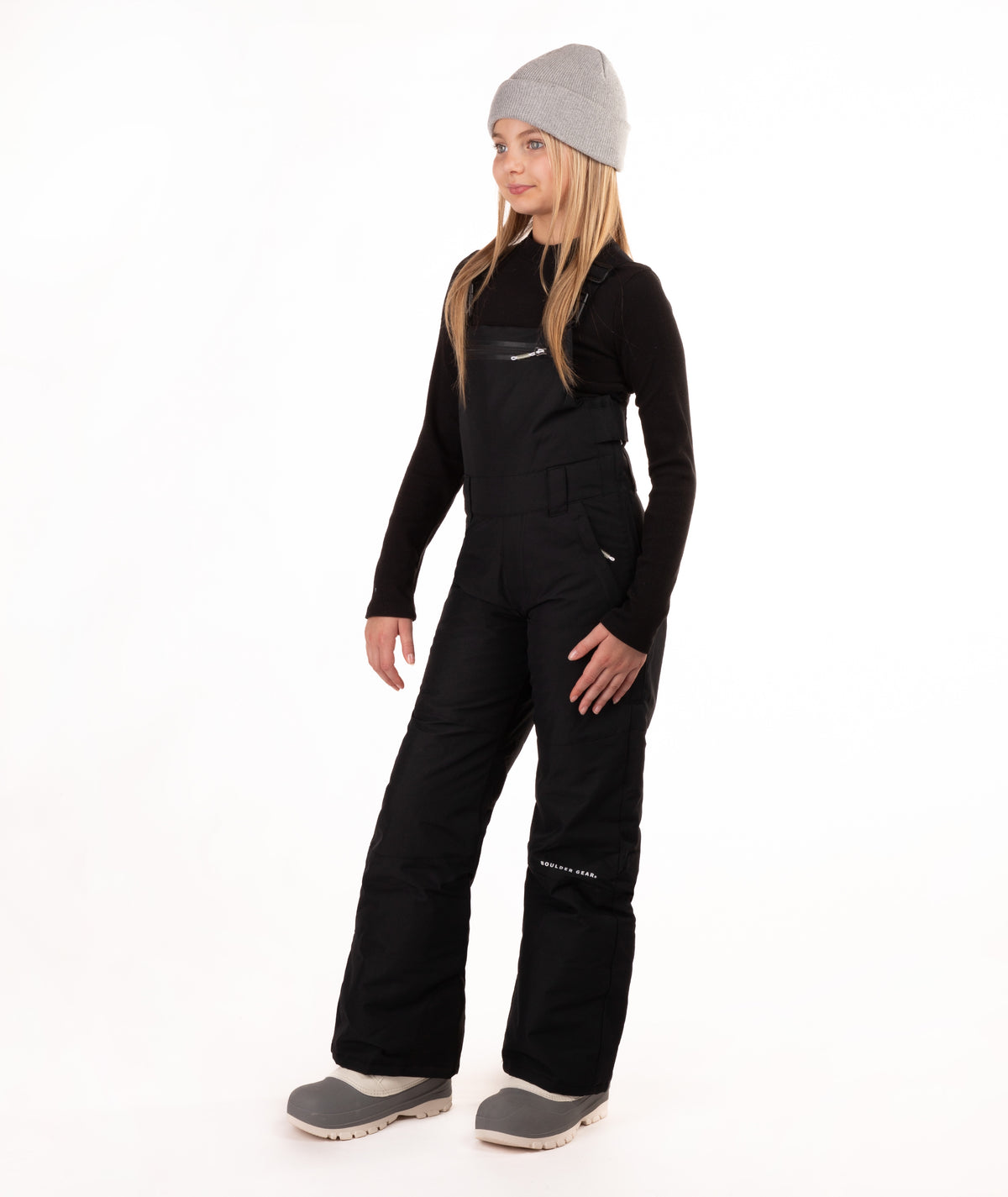 A young girl modeling black snow bib overalls with a gray beanie, black long-sleeve shirt, and gray boots against a white studio background.