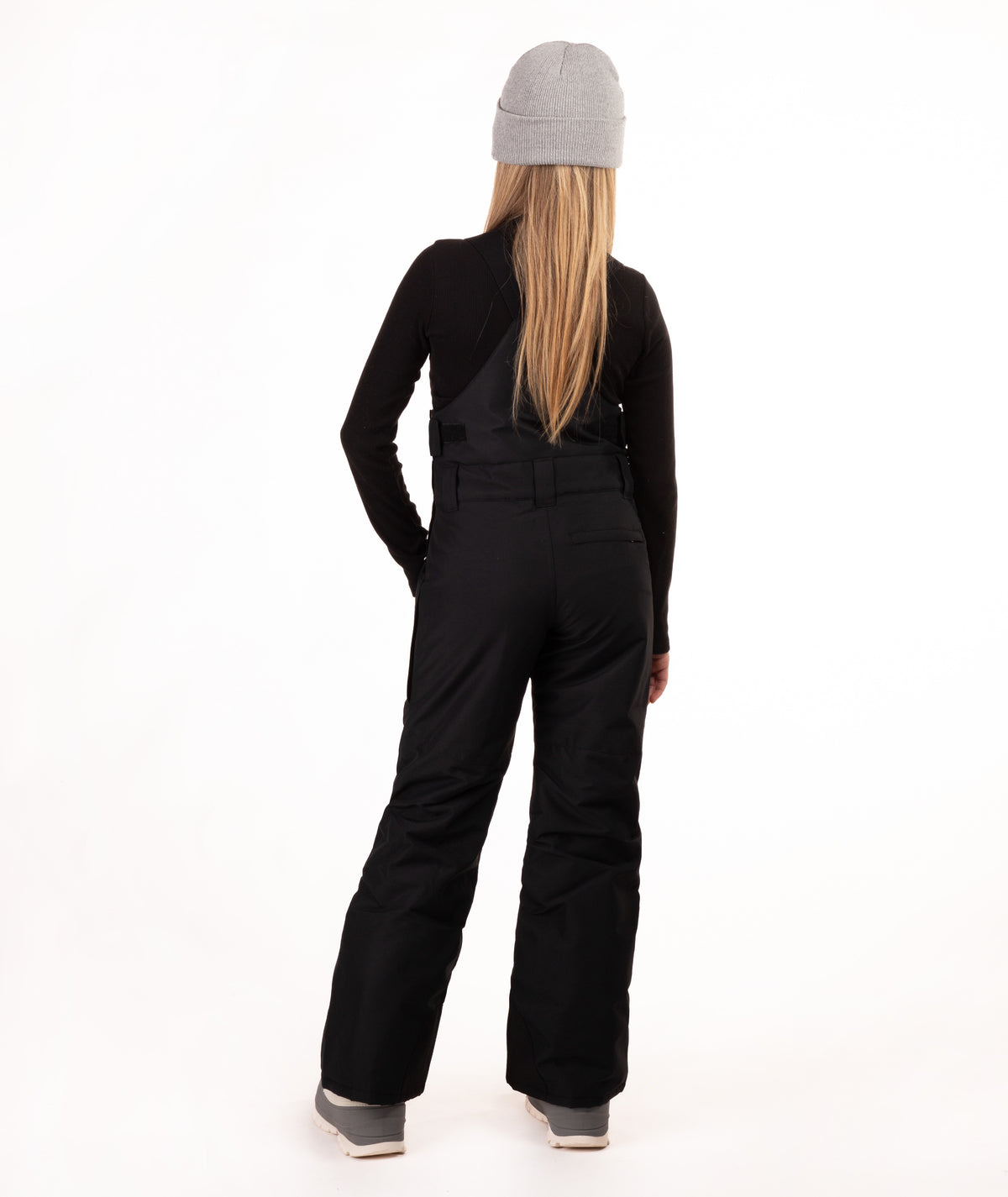 A model shown from the back wearing a black long-sleeve top and black snow bibs, paired with a gray knit beanie and gray boots, against a solid white studio background.