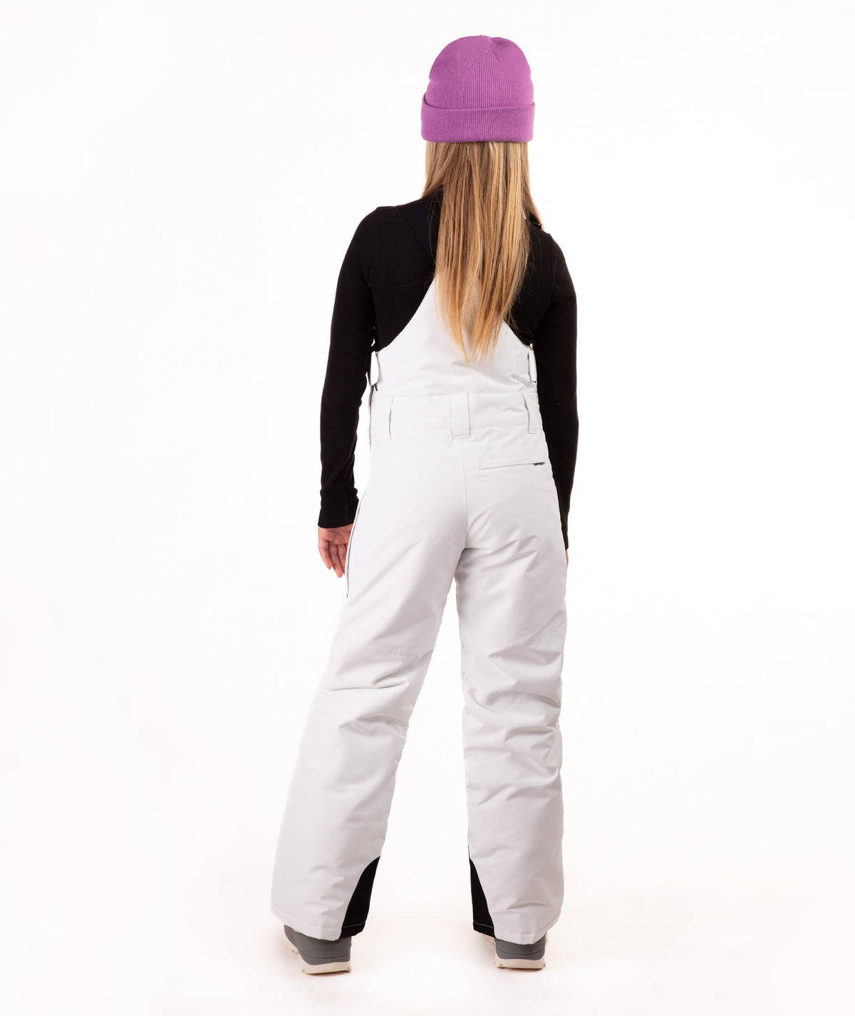 A model viewed from the back wearing white snow bibs, a black long-sleeve top, a purple beanie, and gray boots, set against a plain white studio background.