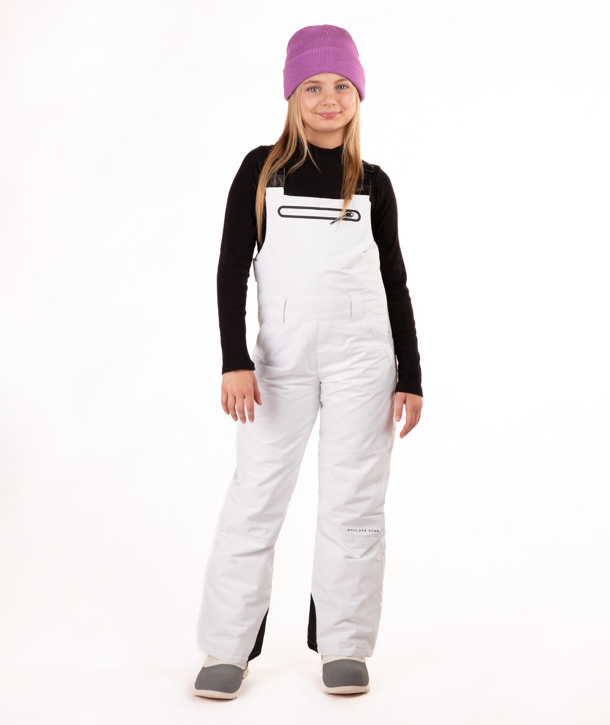 A young girl modeling white snow bib overalls, a black long-sleeve shirt, gray boots, and a purple knit beanie, standing against a solid white background.