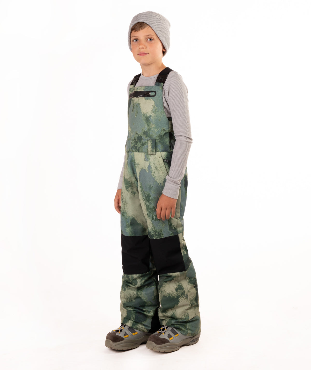 A child wearing camouflage snow bib overalls with black knee panels, a gray beanie, a light gray long-sleeve shirt, and boots, standing against a white studio background.