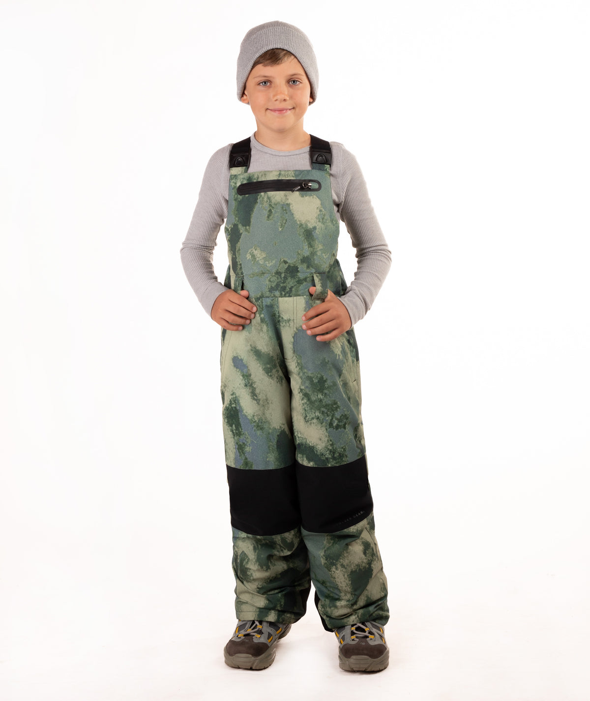A child wearing camouflage snow bib overalls with black knee panels, a gray beanie, a light gray long-sleeve shirt, and boots, standing against a white studio background.