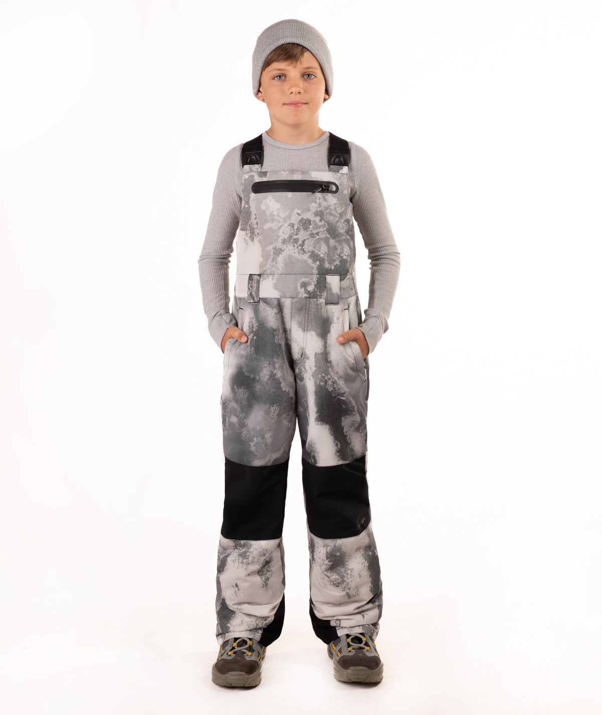 A child viewed from the back wearing gray and white tie-dye snow bib overalls, a gray long-sleeve shirt, and a gray beanie on a white studio background.