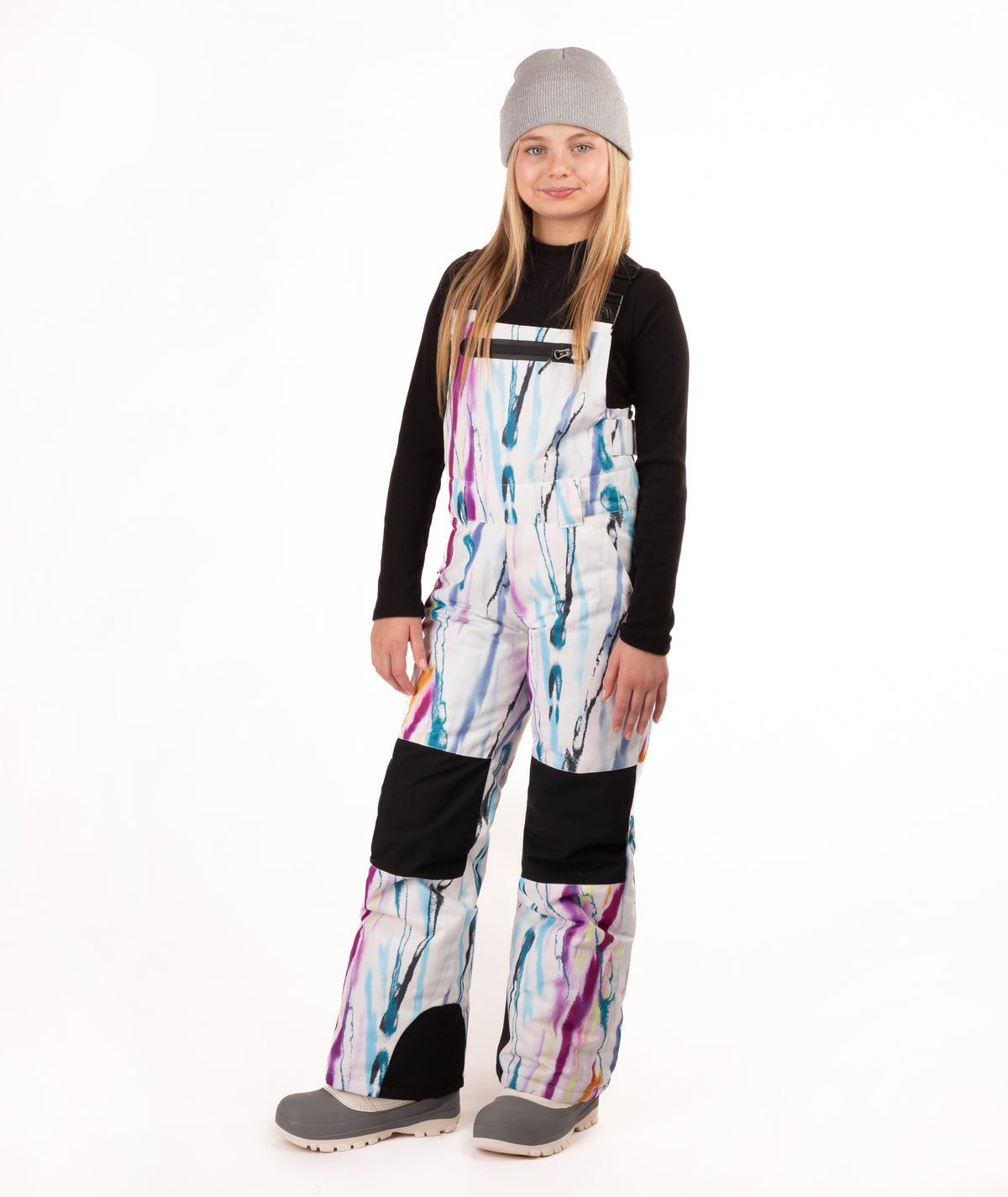 A young girl wearing a black long-sleeve shirt and colorful snow bib overalls with a white base and purple, blue, and pink paint-stroke pattern, along with a gray beanie and gray shoes, standing against a white studio background.