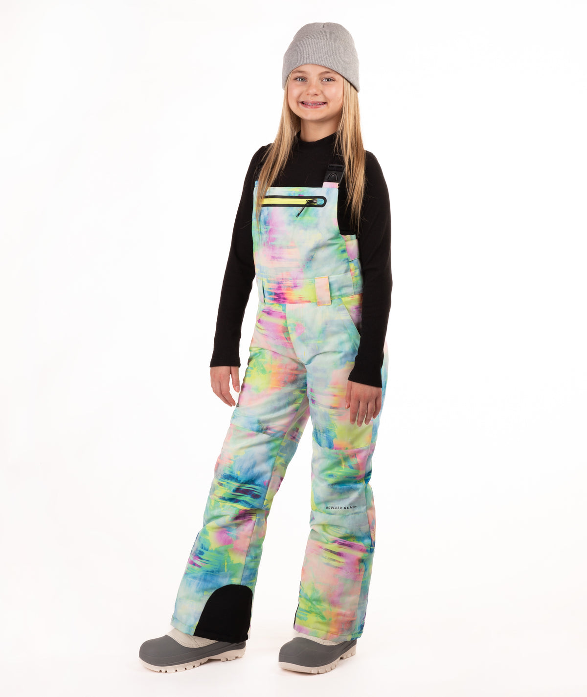 A girl wearing a gray beanie, black long-sleeve shirt, and colorful pastel tie-dye snow bib overalls with gray boots, standing against a plain white studio background.