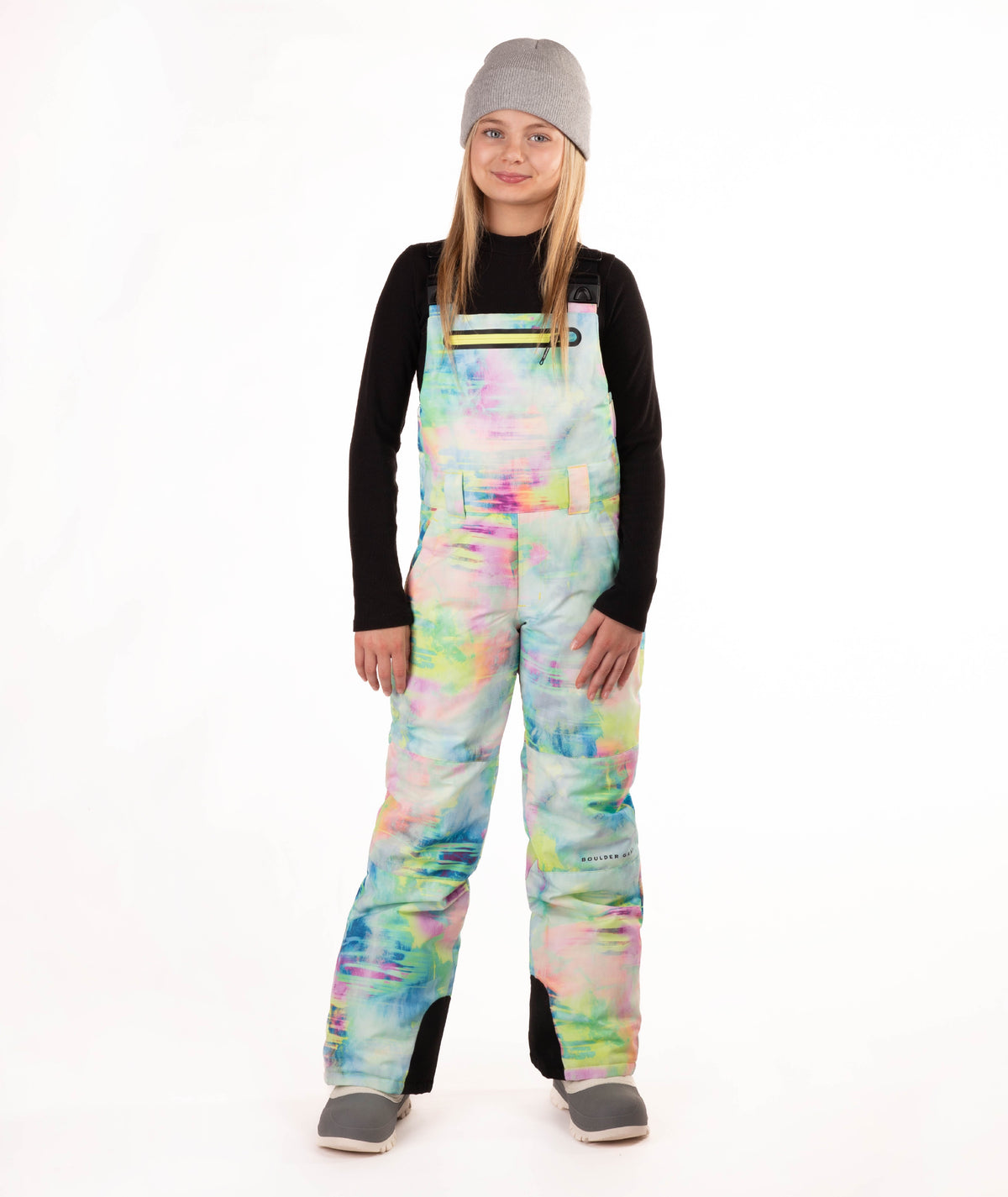 A girl wearing a gray beanie, black long-sleeve shirt, and colorful pastel tie-dye snow bib overalls with gray boots, standing against a plain white studio background.